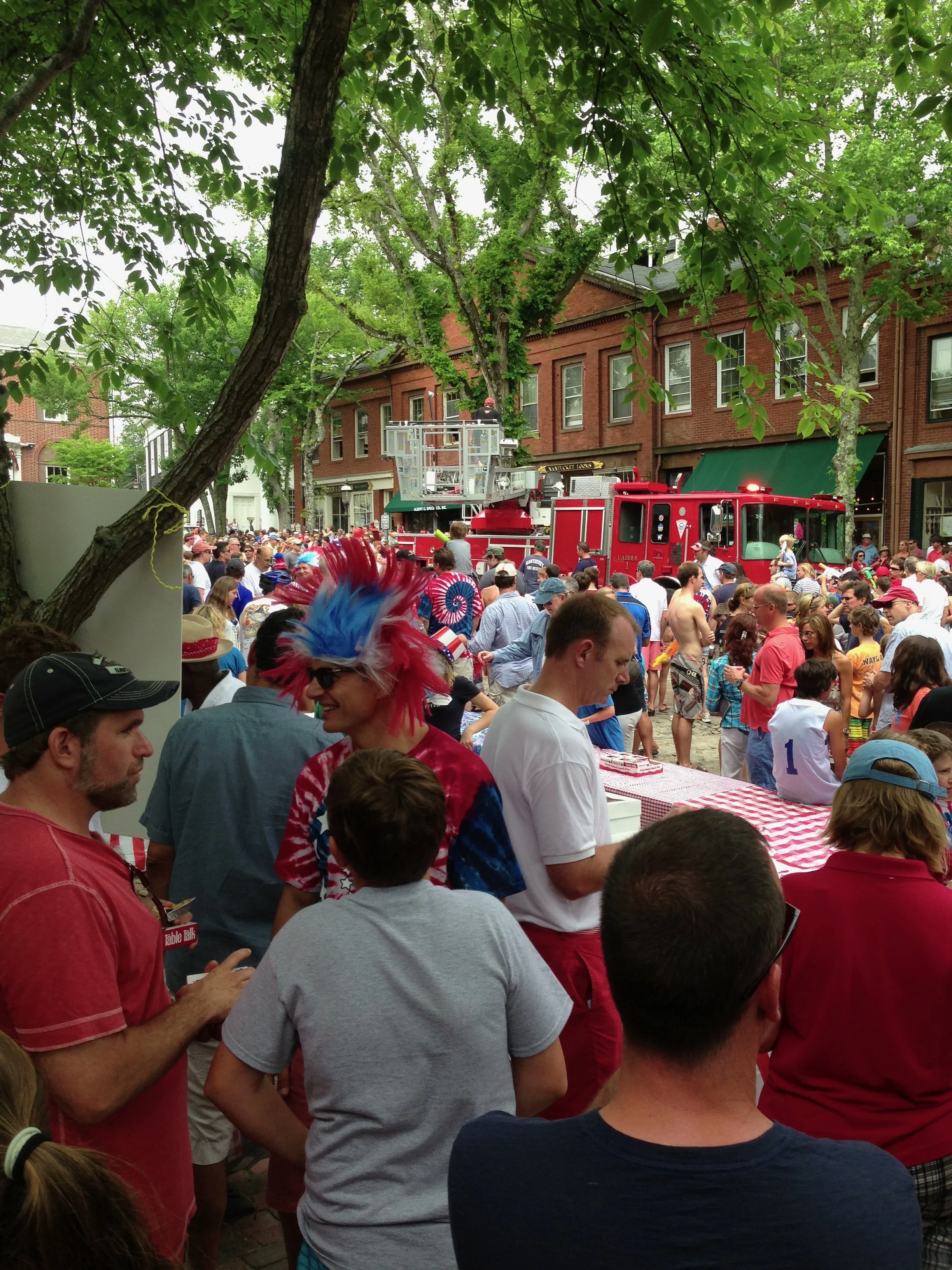 Large crowd gathered outdoors during a festive event with a fire truck and a person in a red, white, and blue wig.