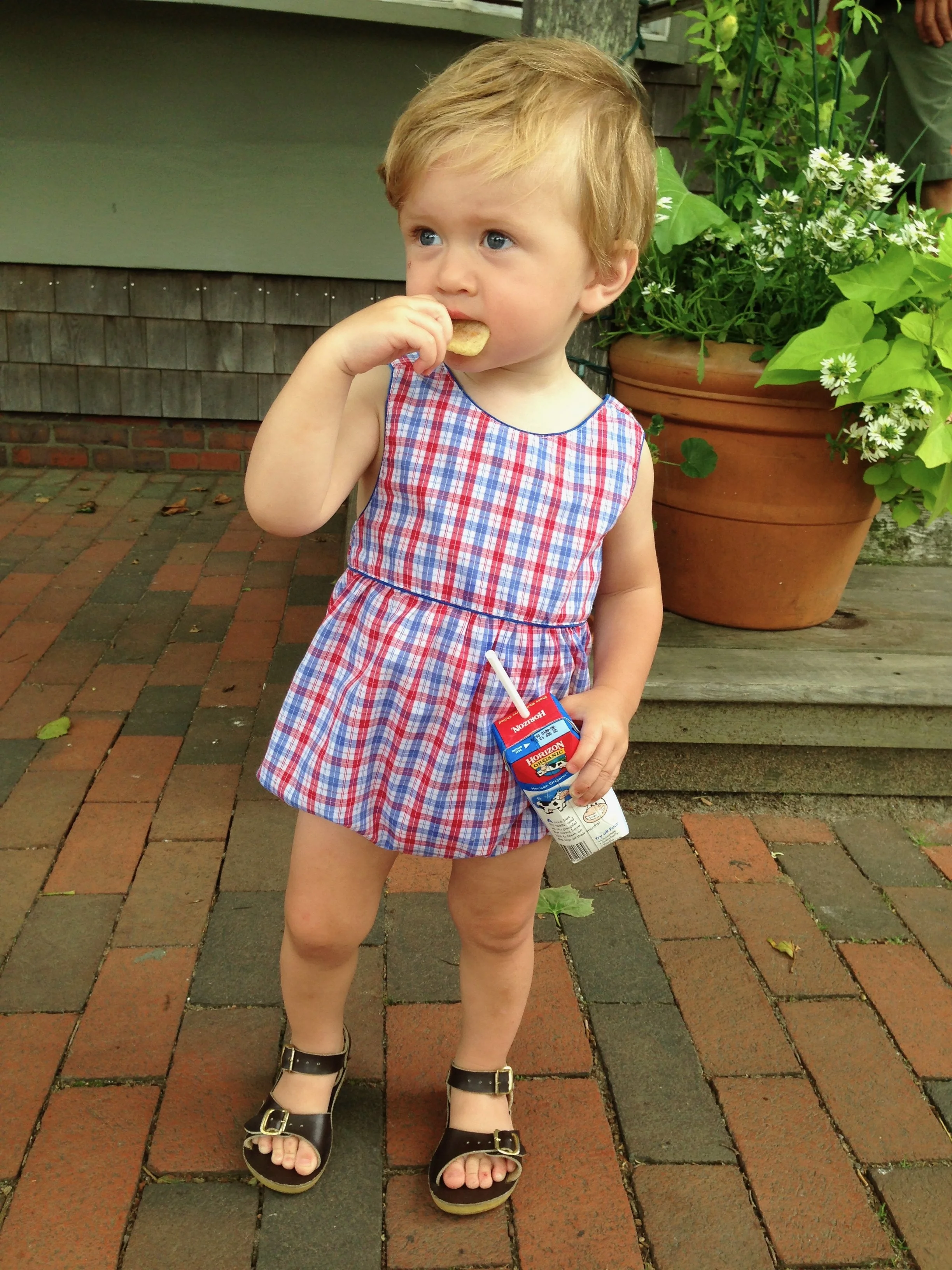 A young boy in a red, white, and blue plaid dress, wearing sandals, standing on a brick patio holding a juice box with a straw and eating a cookie.