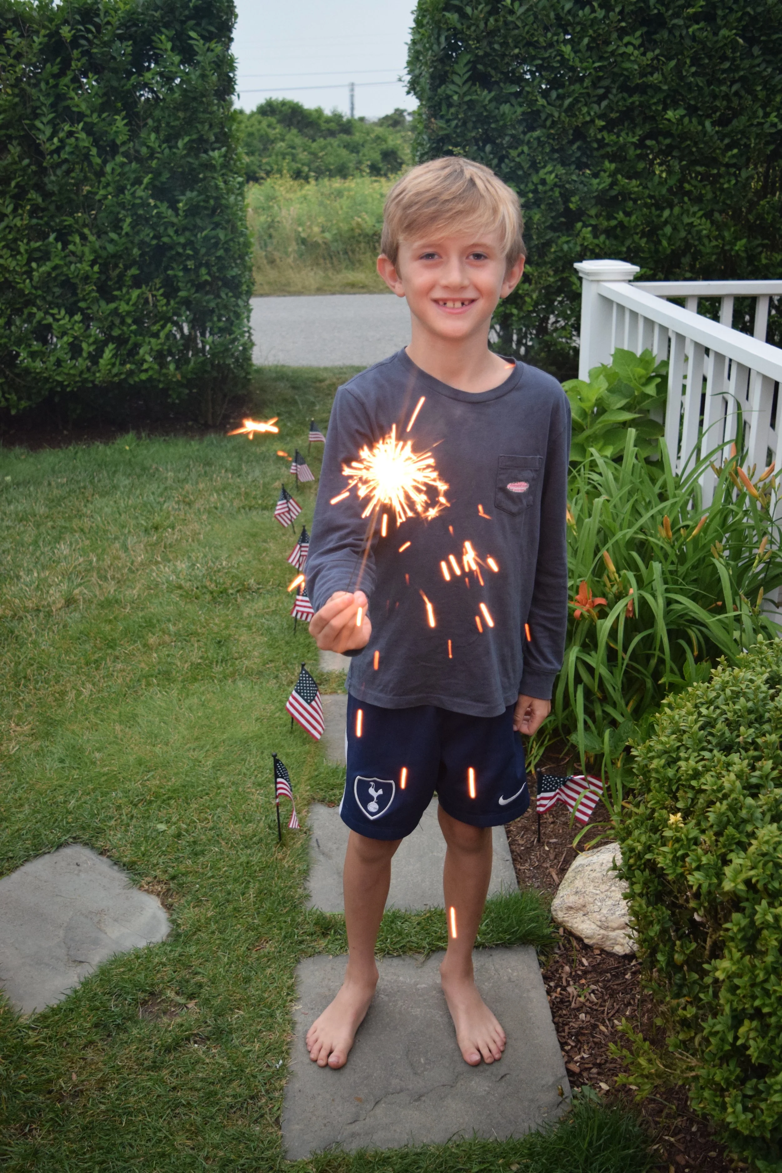 A young boy stands outdoors holding a lit sparkler, smiling. The yard is decorated with small American flags, and the boy is dressed in a long sleeve shirt and shorts, standing barefoot on a stone path surrounded by greenery.