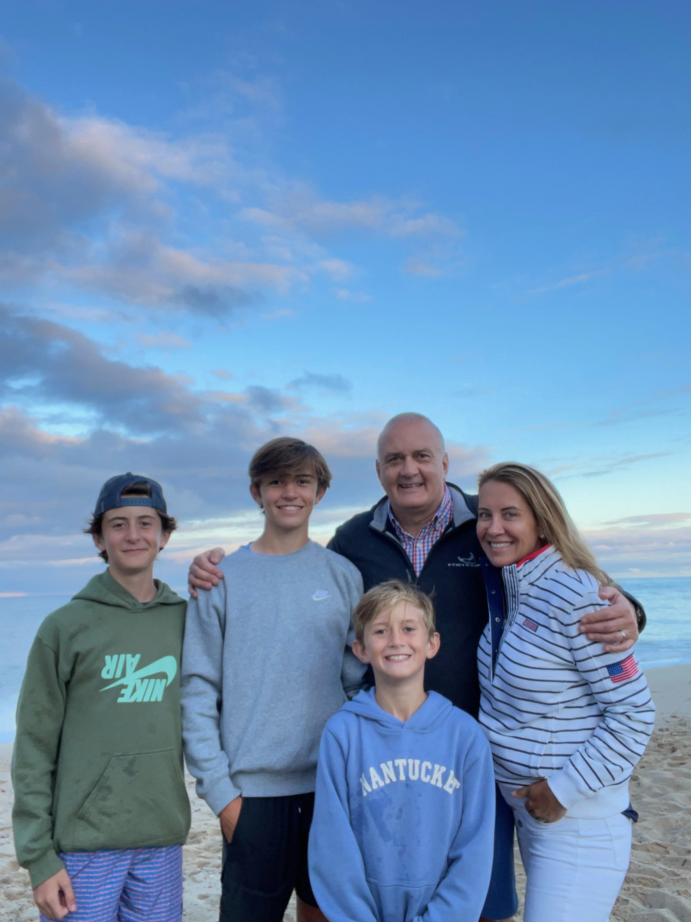 Family of five enjoying time together on the beach with blue sky and clouds in the background, smiling and embracing each other.