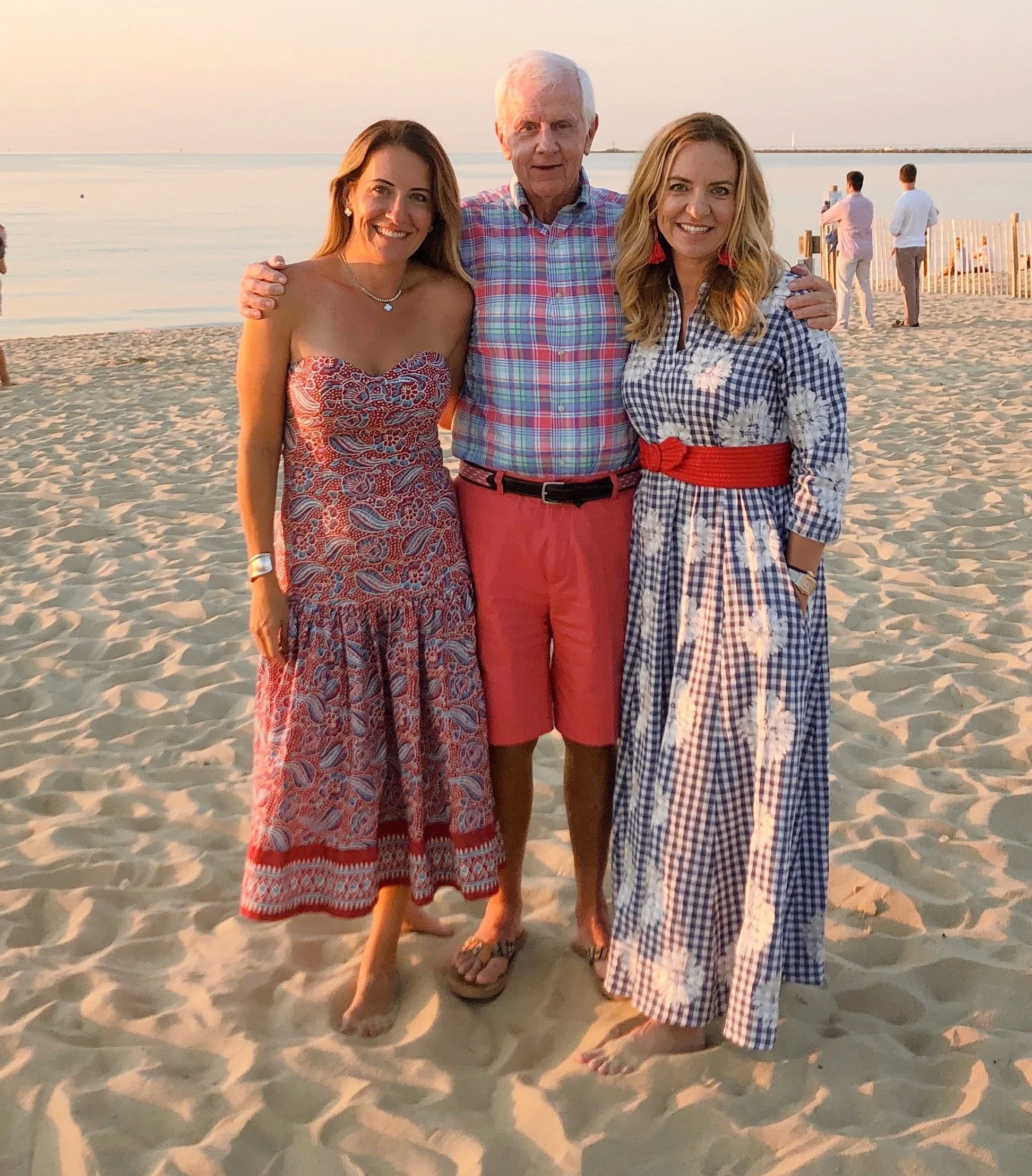 Three people standing on a sandy beach at sunset, smiling, with water and a pier in the background. Two women and one man, dressed in summer and beachwear.