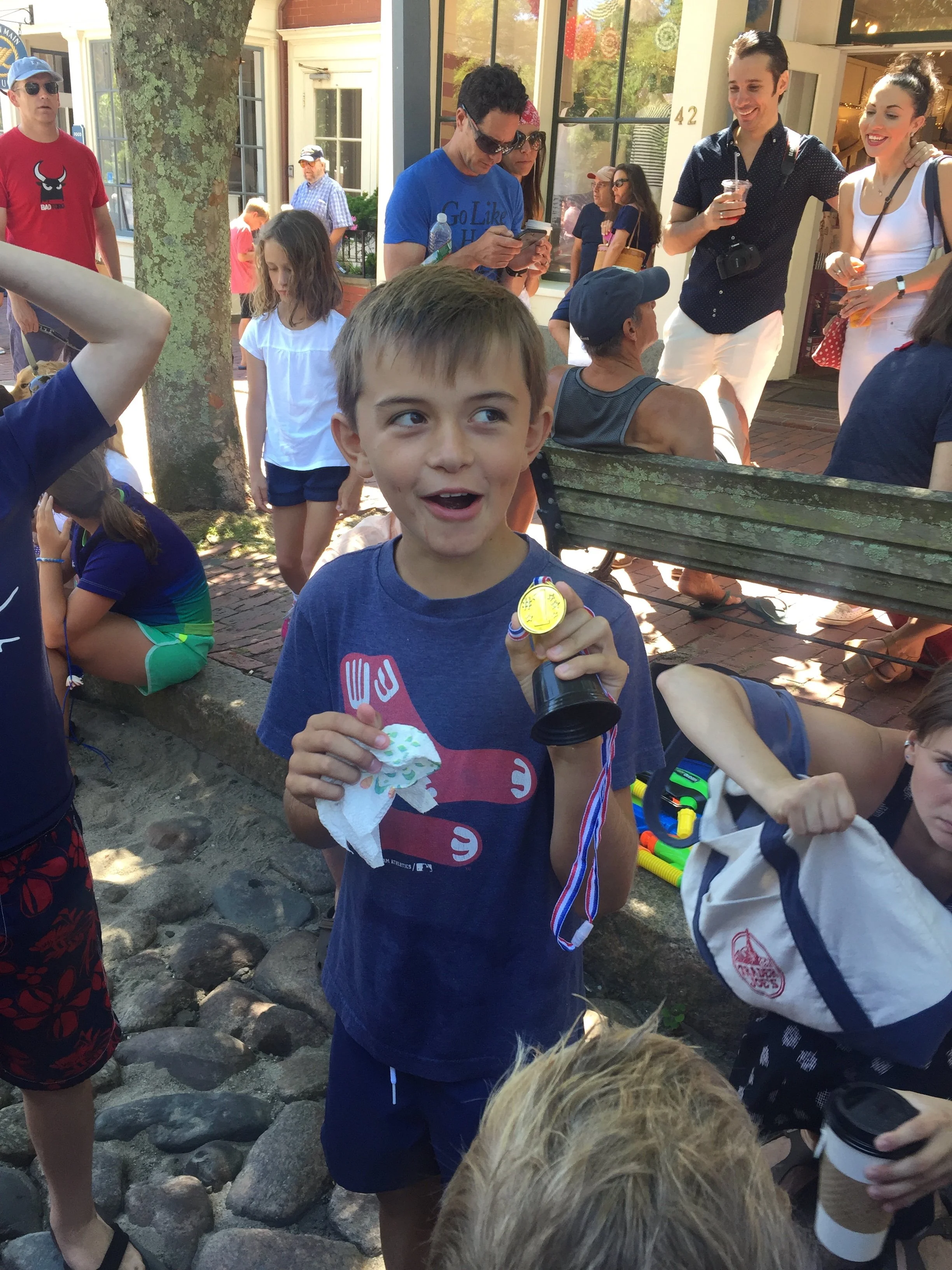 A young boy holding a gold medal and a pacifier, smiling outdoors at a lively gathering of children and adults.