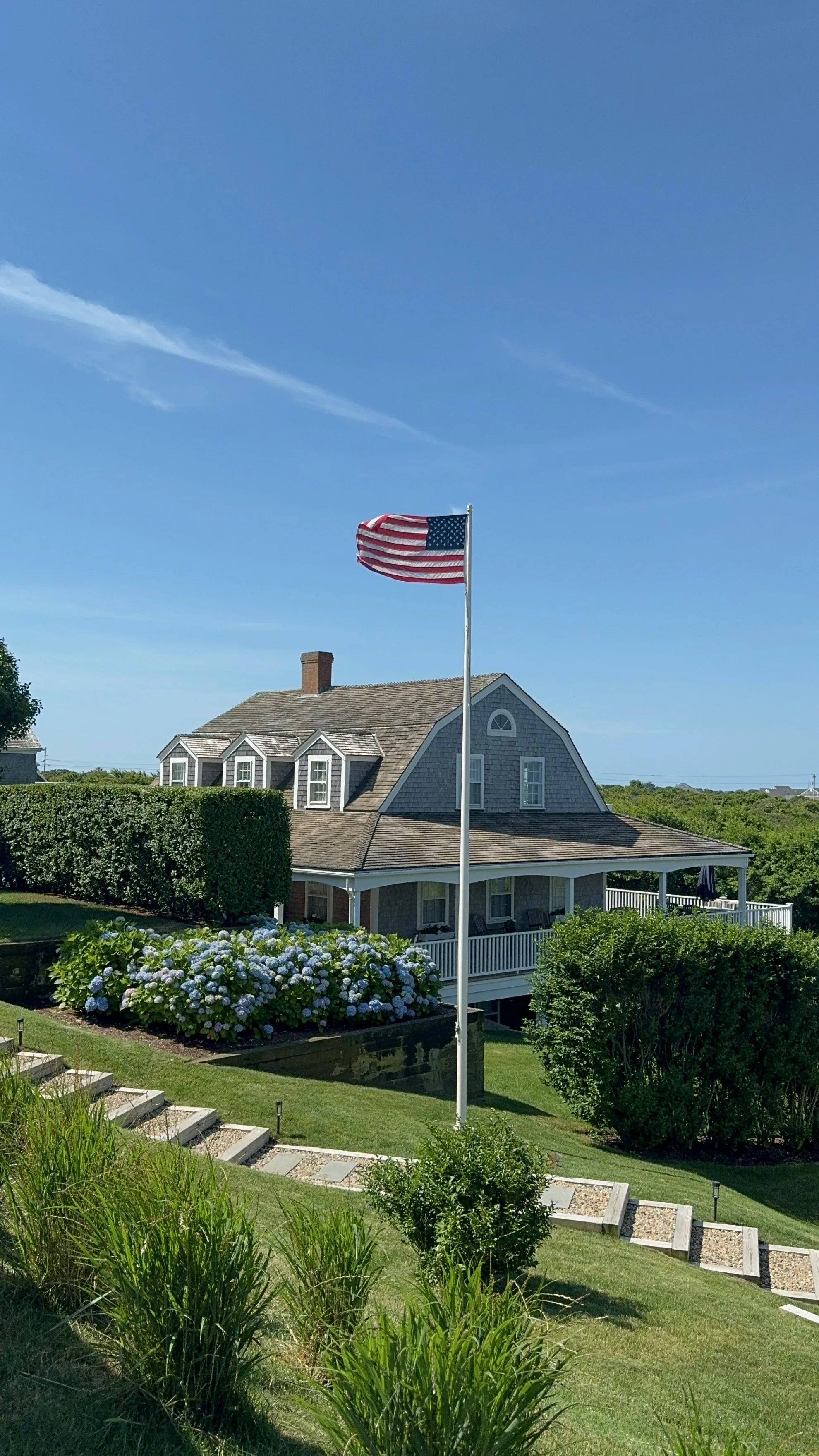 A house with a porch and a landscaped yard, featuring an American flag waving on a tall flagpole under a clear blue sky.