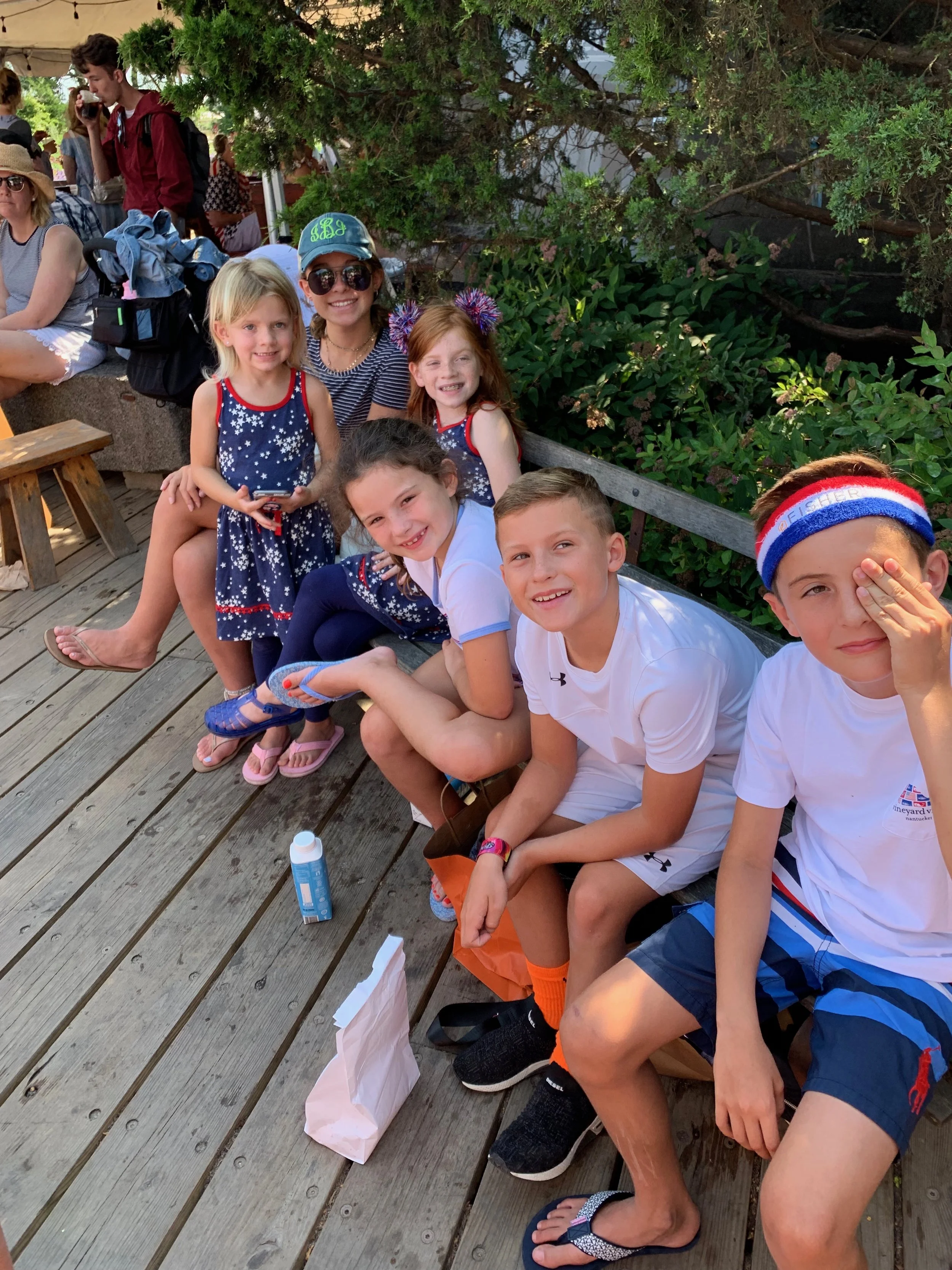 Group of children and a woman sitting and standing on a wooden deck outdoors, with trees and bushes in the background, enjoying a sunny day.