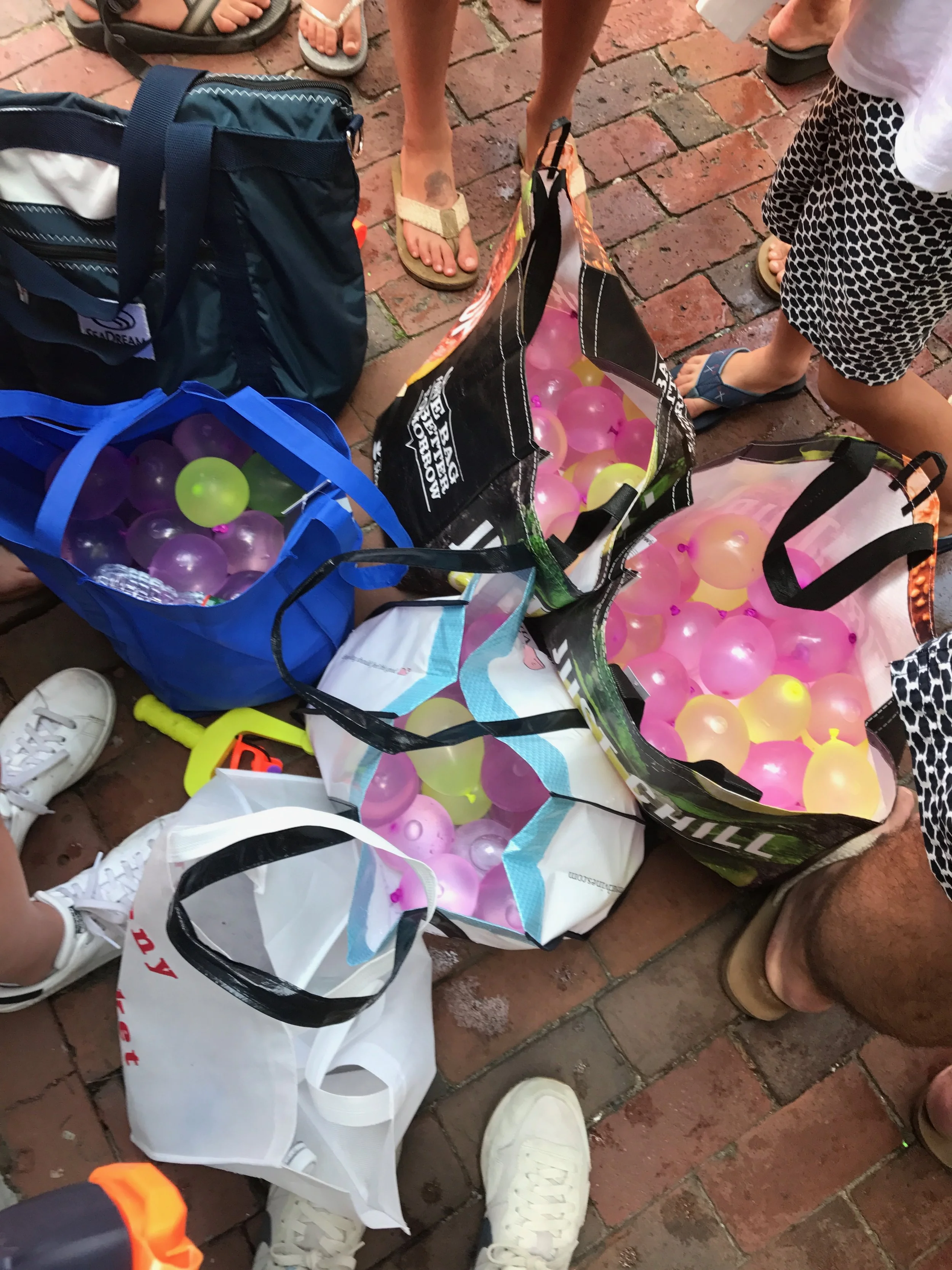 Several bags on a brick ground filled with colorful plastic balls, surrounded by people wearing sandals and sneakers.