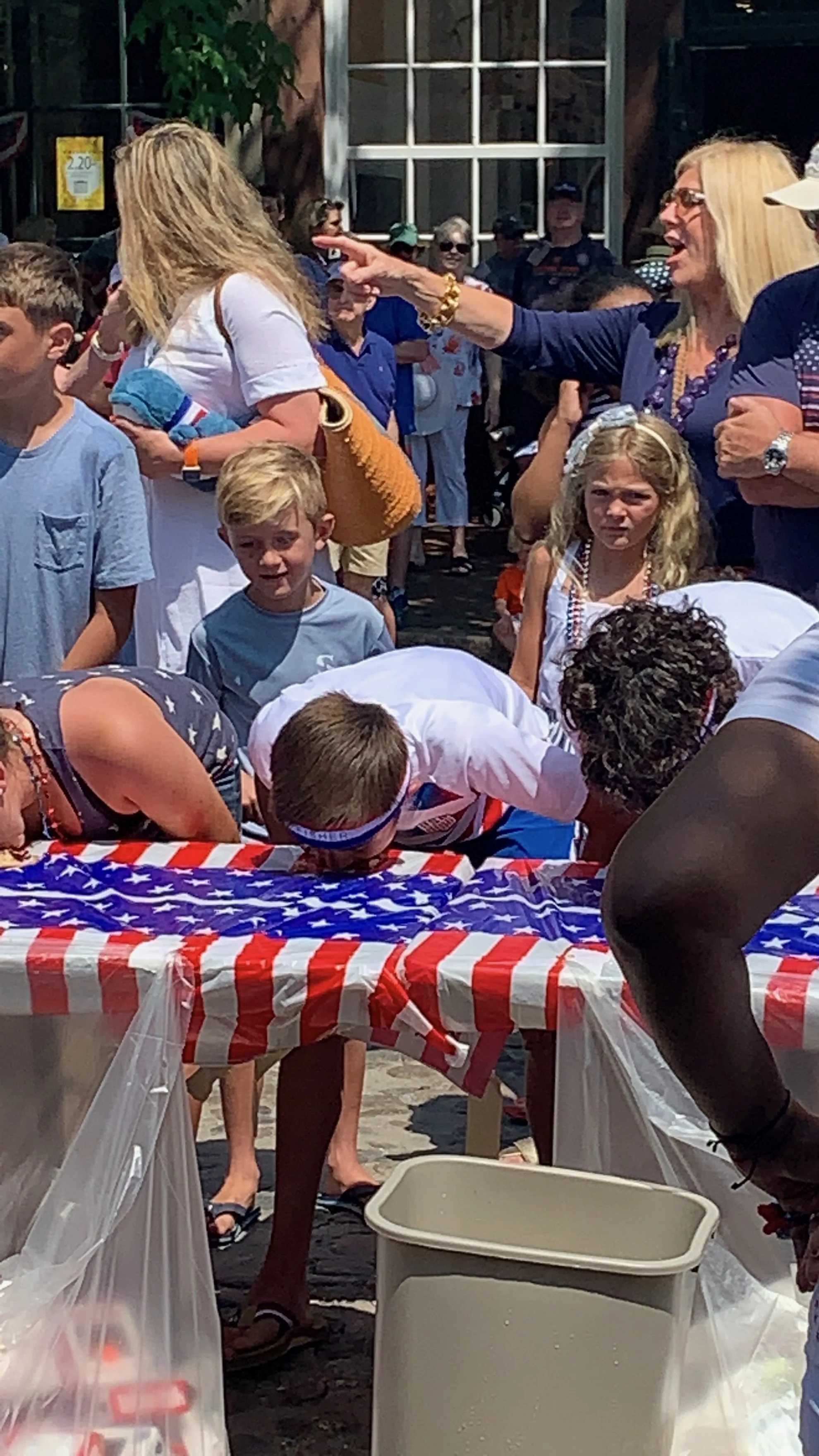 People participating in an outdoor event, with some leaning over a table covered with an American flag-themed tablecloth, possibly during a game or activity, with onlookers and a woman pointing in the background.