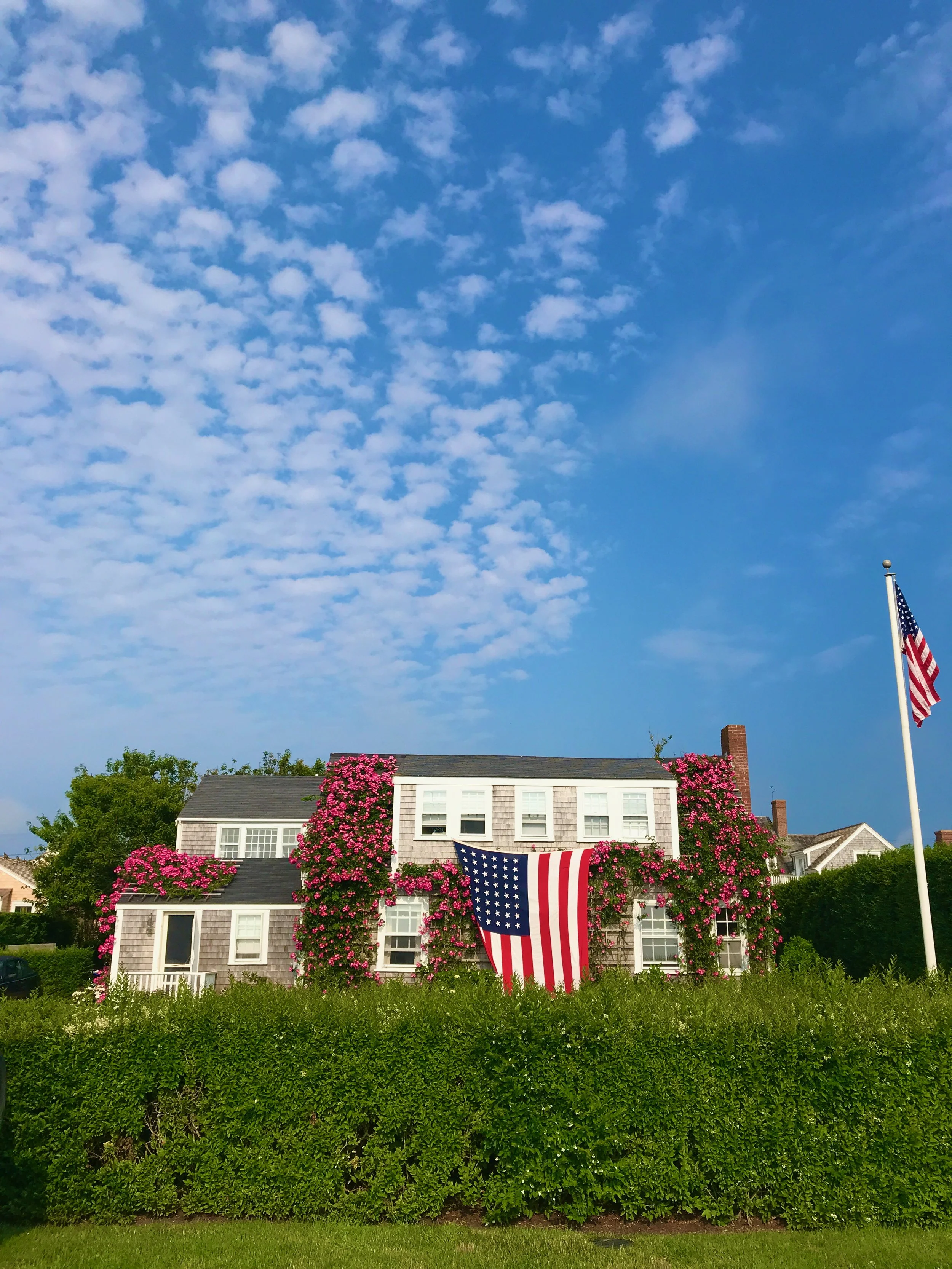 A house decorated with pink flowers and two American flags, one large hanging on the house and one on a flagpole to the right, against a blue sky with scattered clouds.