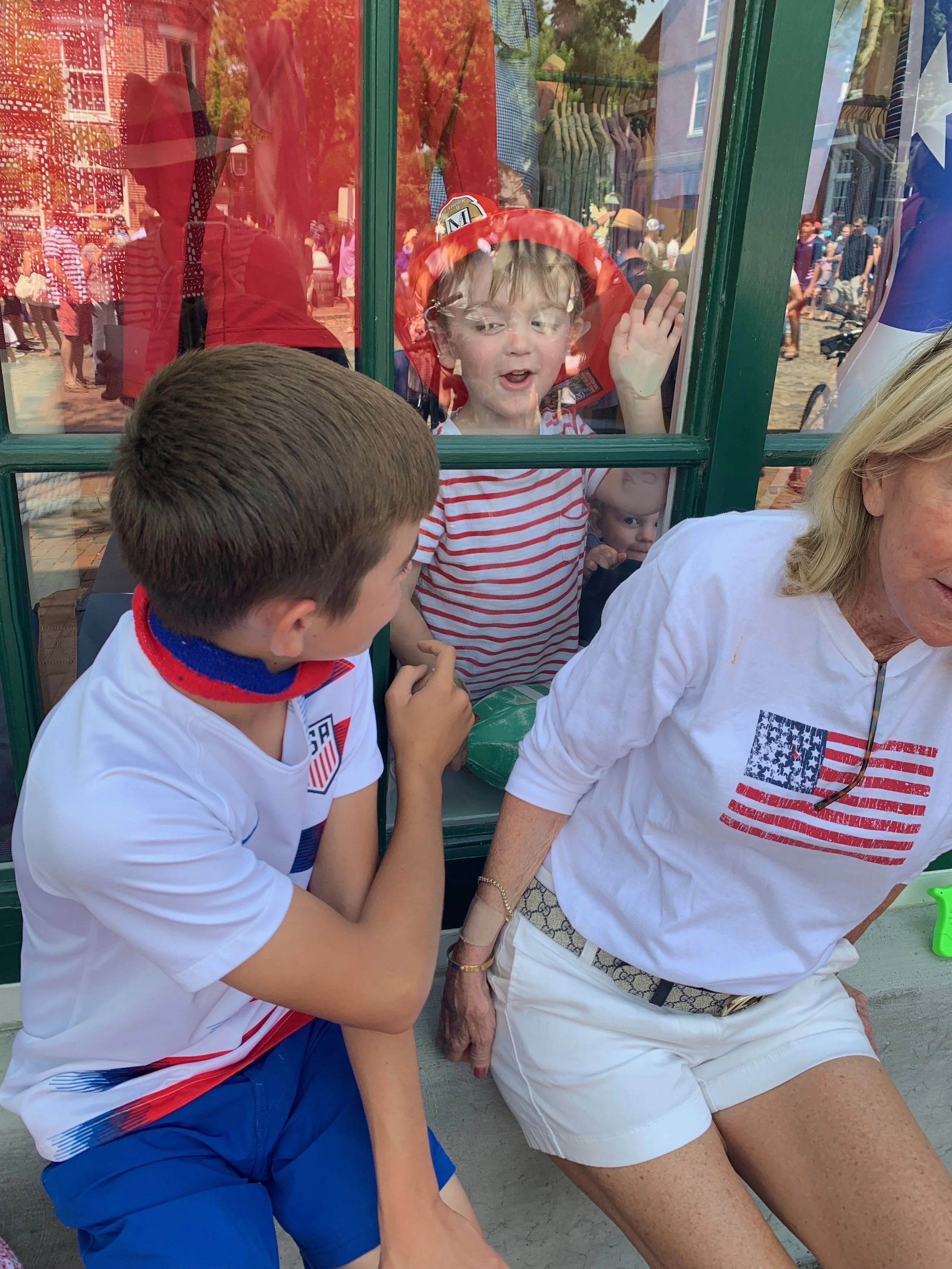 A young boy and an older woman sit outside at a table, wearing patriotic clothing with American flags. A young child with a firefighter helmet and a red and white striped shirt presses a hand and face against a glass window, looking at the boy and wo