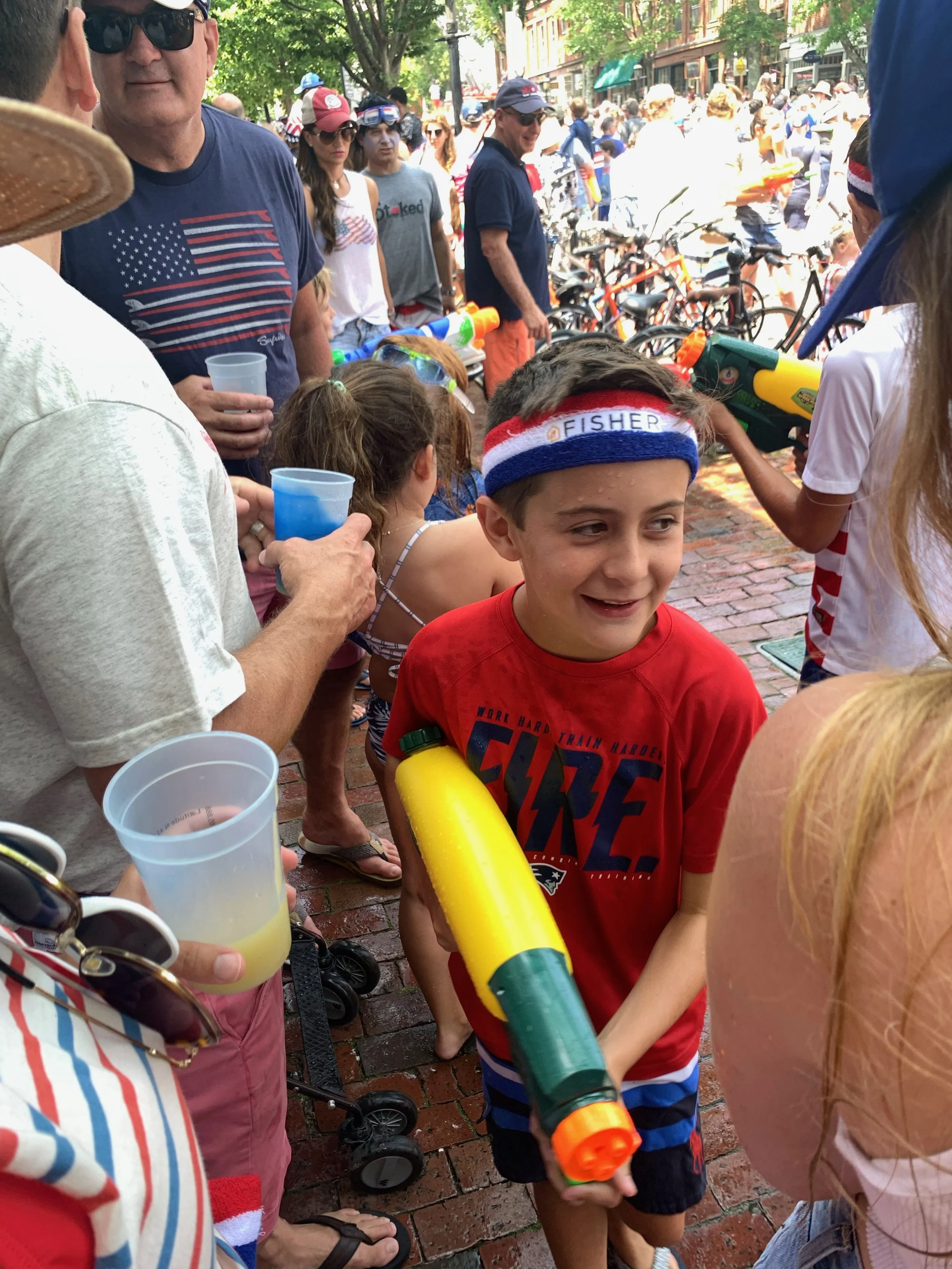 Children and adults participating in a water fight on a brick sidewalk on a sunny day, with water guns and water balloons.