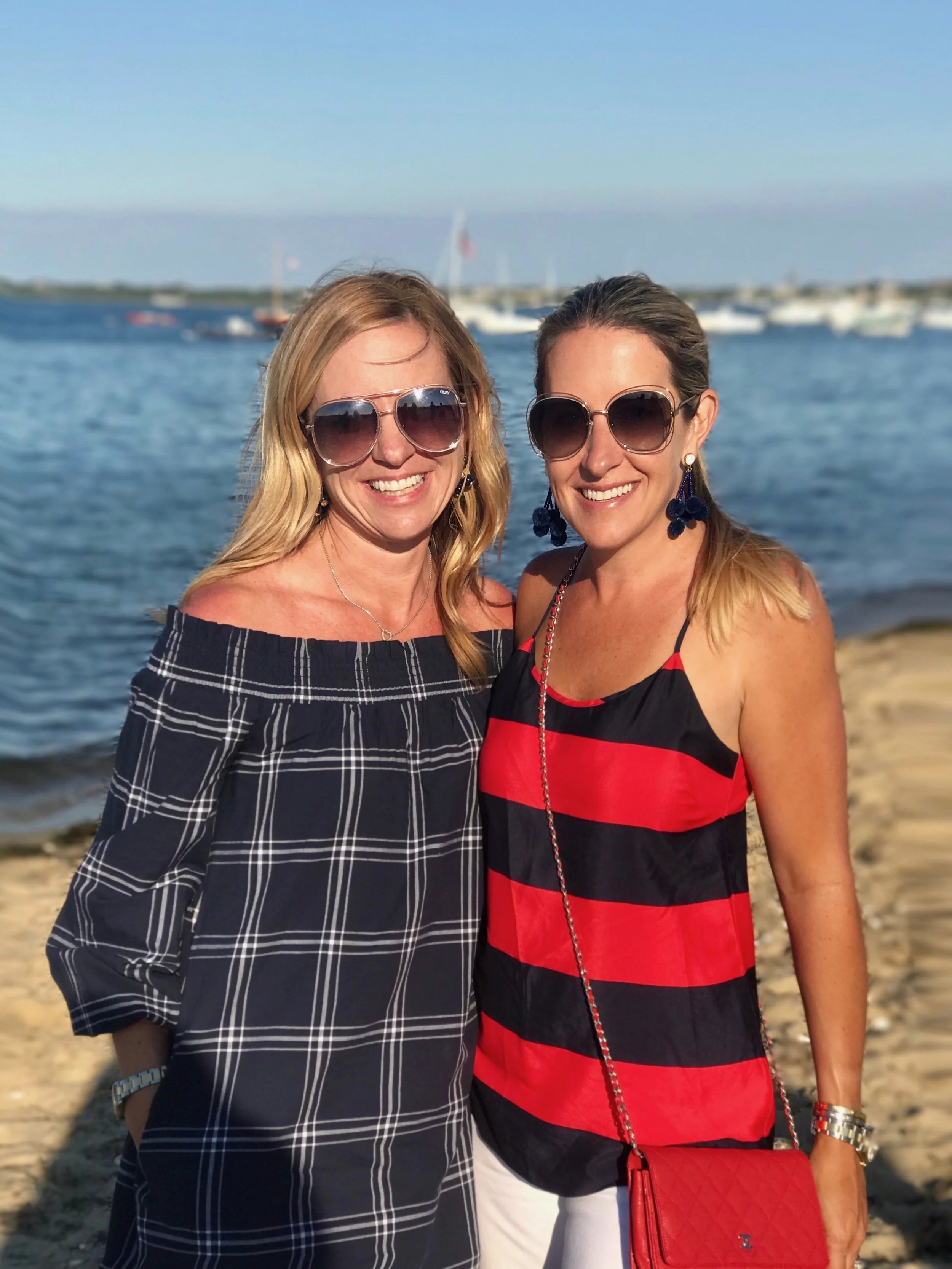 Two women standing on a beach near water, smiling, wearing sunglasses and summer clothes. Boats are visible in the background.