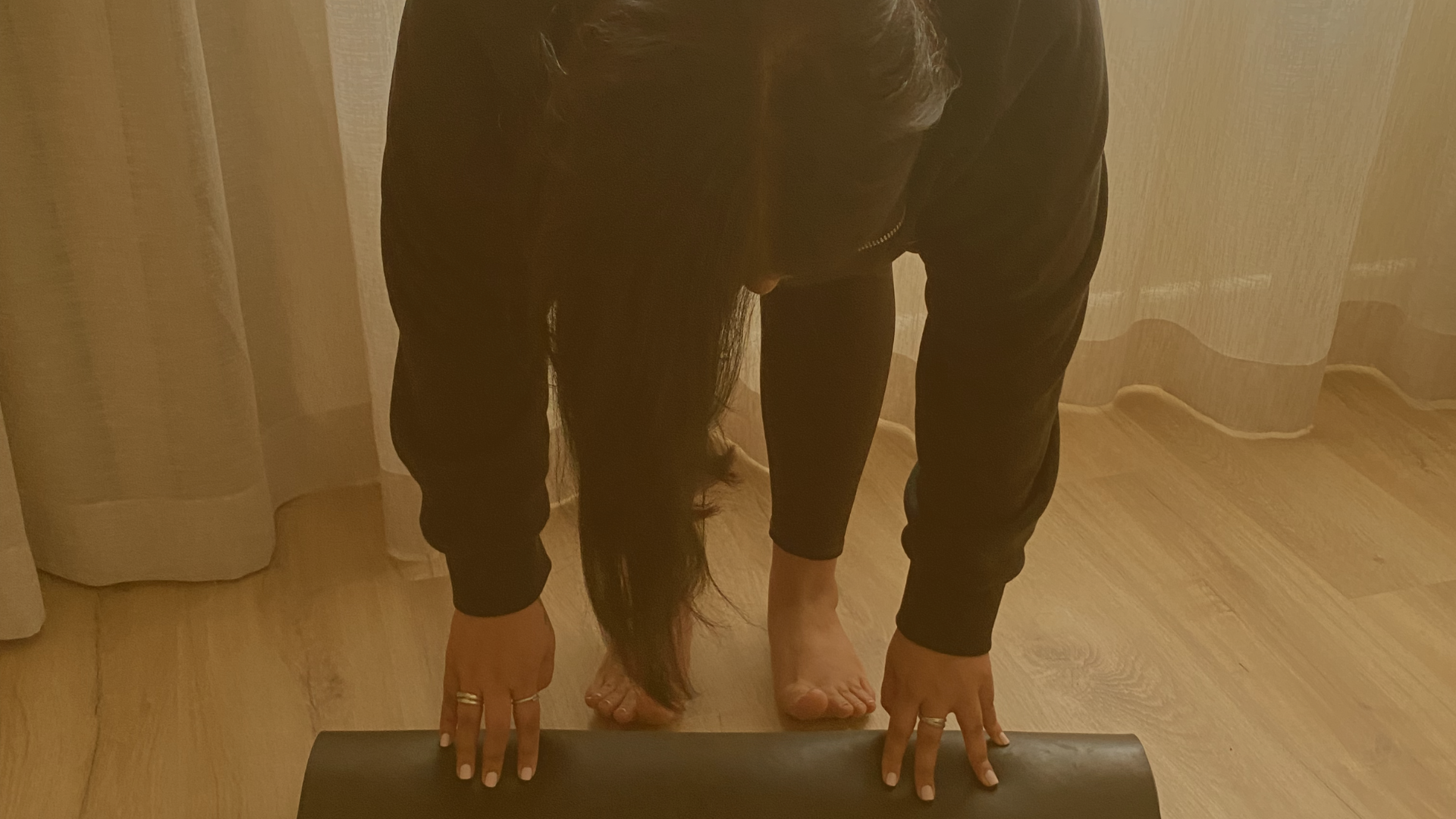Prishaniee practicing yoga or stretching indoors, seen from front, bending over with her hands on the ground and feet flat on the floor, with long dark hair hanging down.
