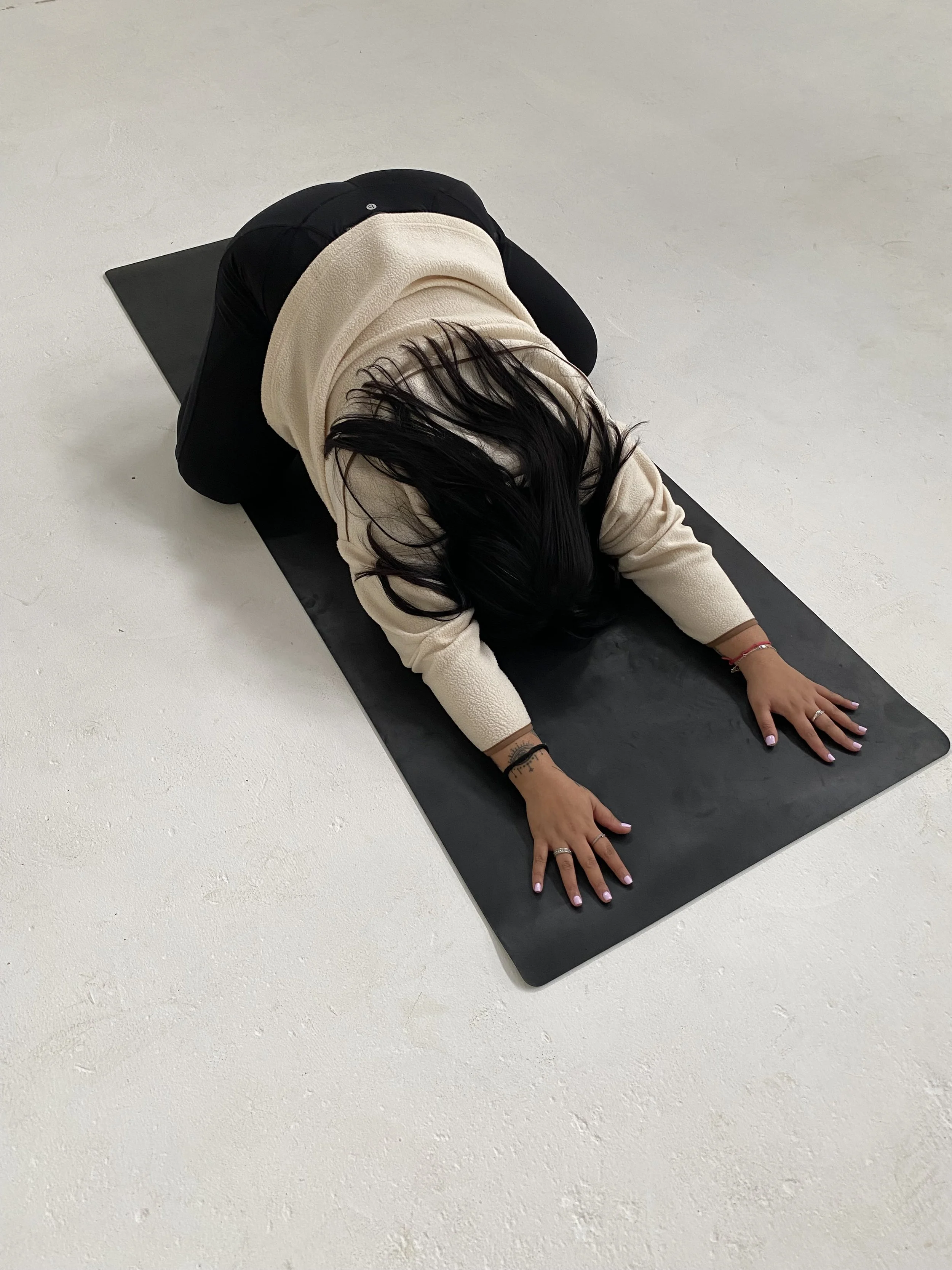 Prishaniee performing child's pose yoga stretch on black mat in a bright, empty room.