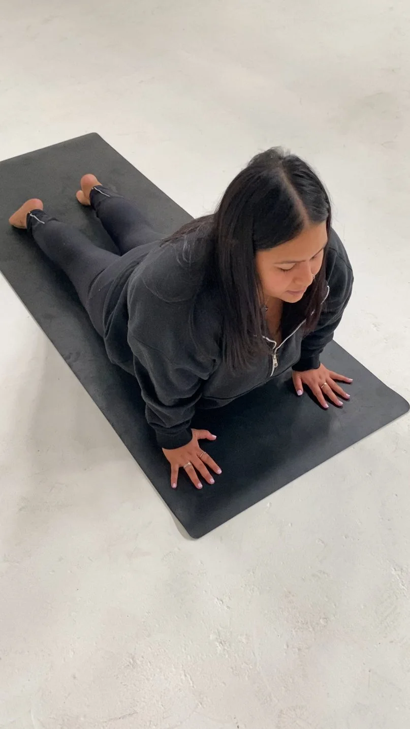 Prishaniee in black hoodie practicing yoga on black mat in a prone position with hands and feet on the ground.