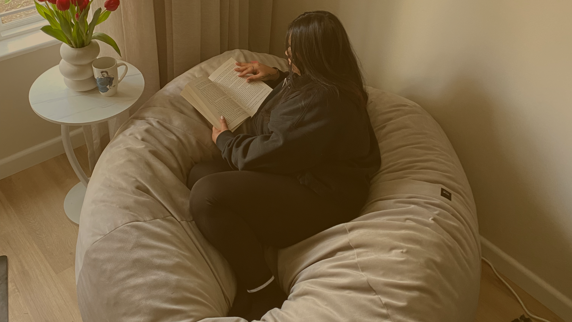 Prishaniee reading a book on a beige, fluffy corner sofa in a cozy living room. A white side table beside her holds a white vase with pink tulips and a mug with a printed photo.