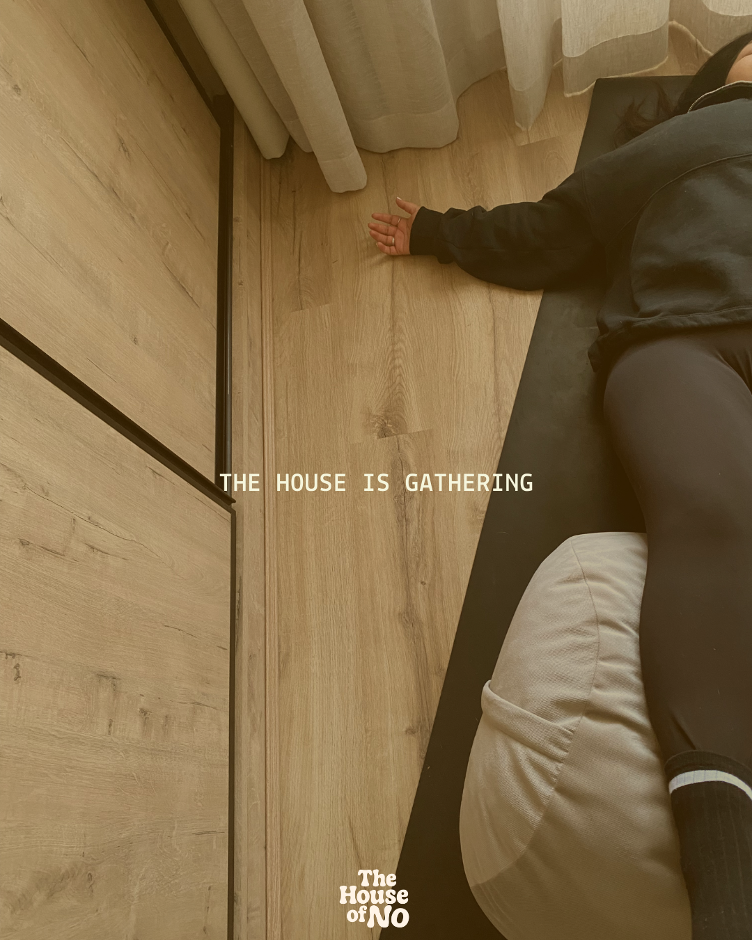 Prishaniee lying on a black couch with a beige cushion, with their arm extended towards a wooden floor and a wooden wall. The scene appears to be in a cozy interior space. Text overlay reads: 'The house is gathering.'