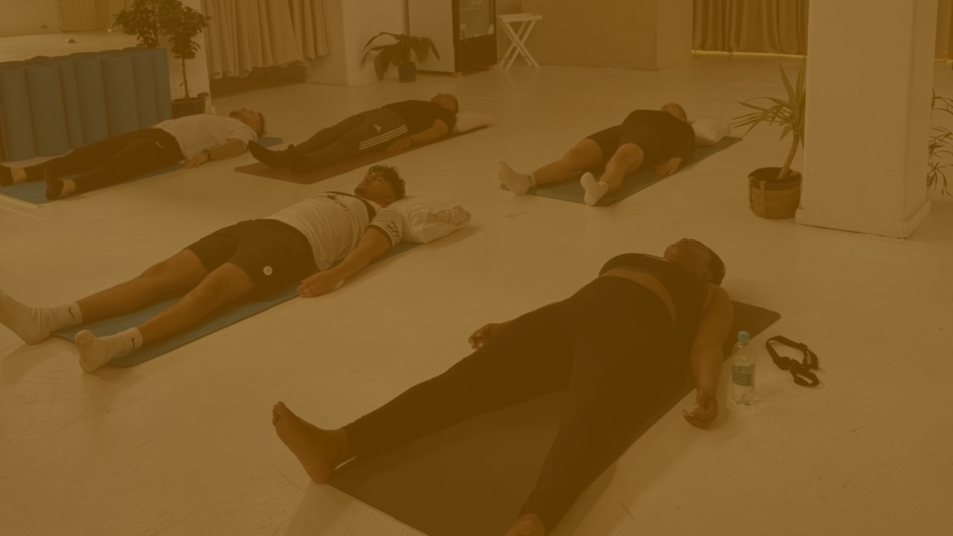 People lying down on mats in a yoga or meditation class, practicing relaxation or meditation in a bright indoor space.