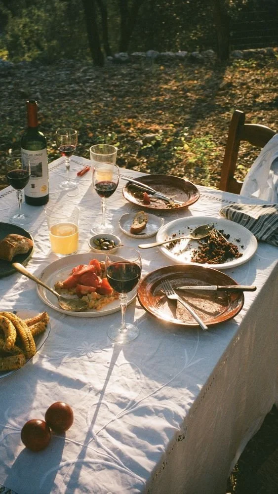 Outdoor dining table with plates, wine glasses, red wine bottle, corn on the cob, tomatoes, and various dishes on a white tablecloth.