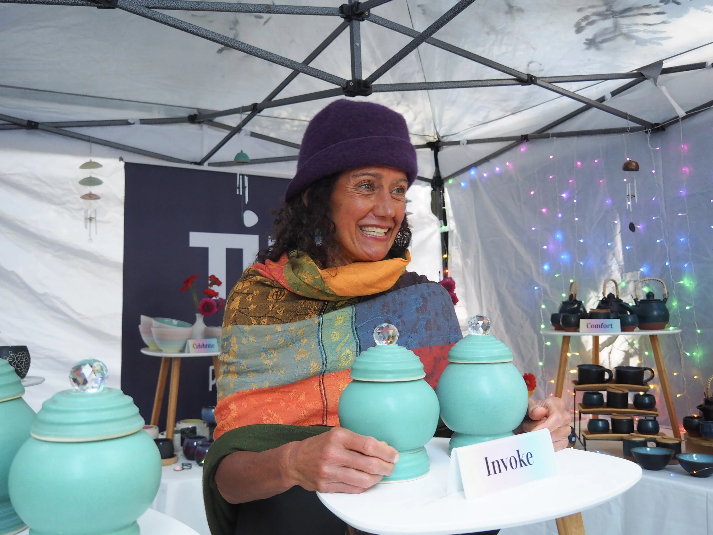 Woman at market stall displaying turquoise ceramic jars with crystal-like knobs, wearing dark green shirt and purple hat, smiling.