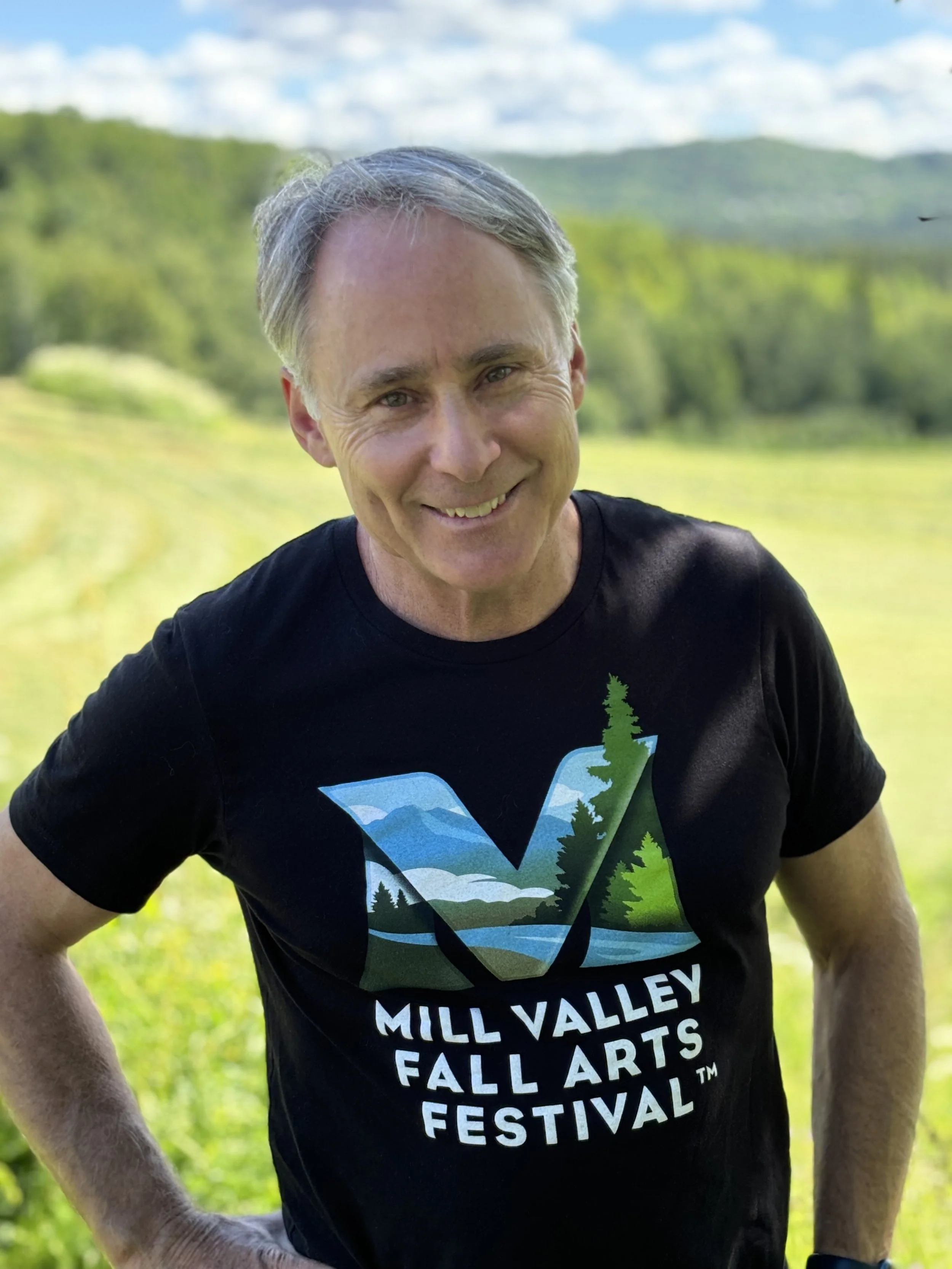 A smiling man outdoors in a field with trees and mountains in the background, wearing a black t-shirt with a nature-themed logo and the text 'Mill Valley Fall Arts Festival'.