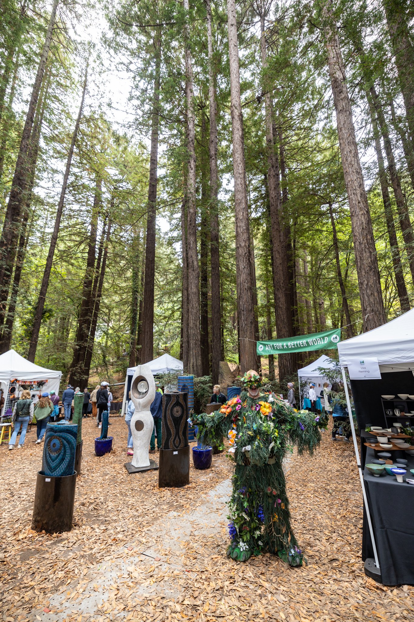 Outdoor art festival in a forest with tall trees, displaying sculptures, pottery, and a person dressed as a floral plant.