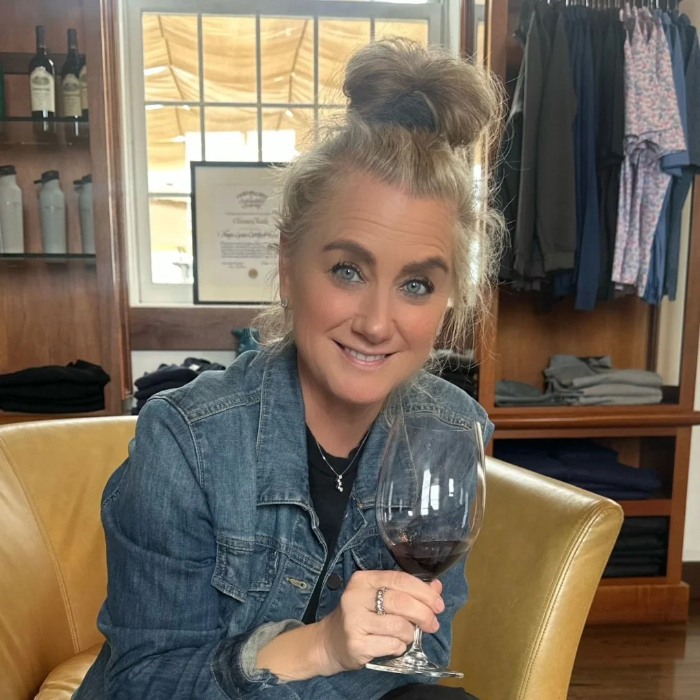 A woman with blue eyes and blond hair in a messy bun, wearing a denim jacket, sitting on a yellow chair, holding a glass of red wine, smiling indoors with wooden shelves and clothing in the background.