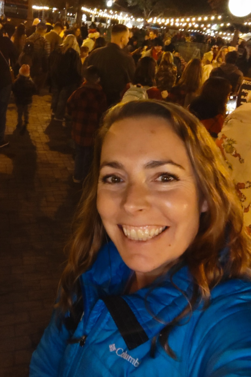A woman with shoulder-length brown hair, smiling, wearing a white top and a necklace, standing against a neutral background.