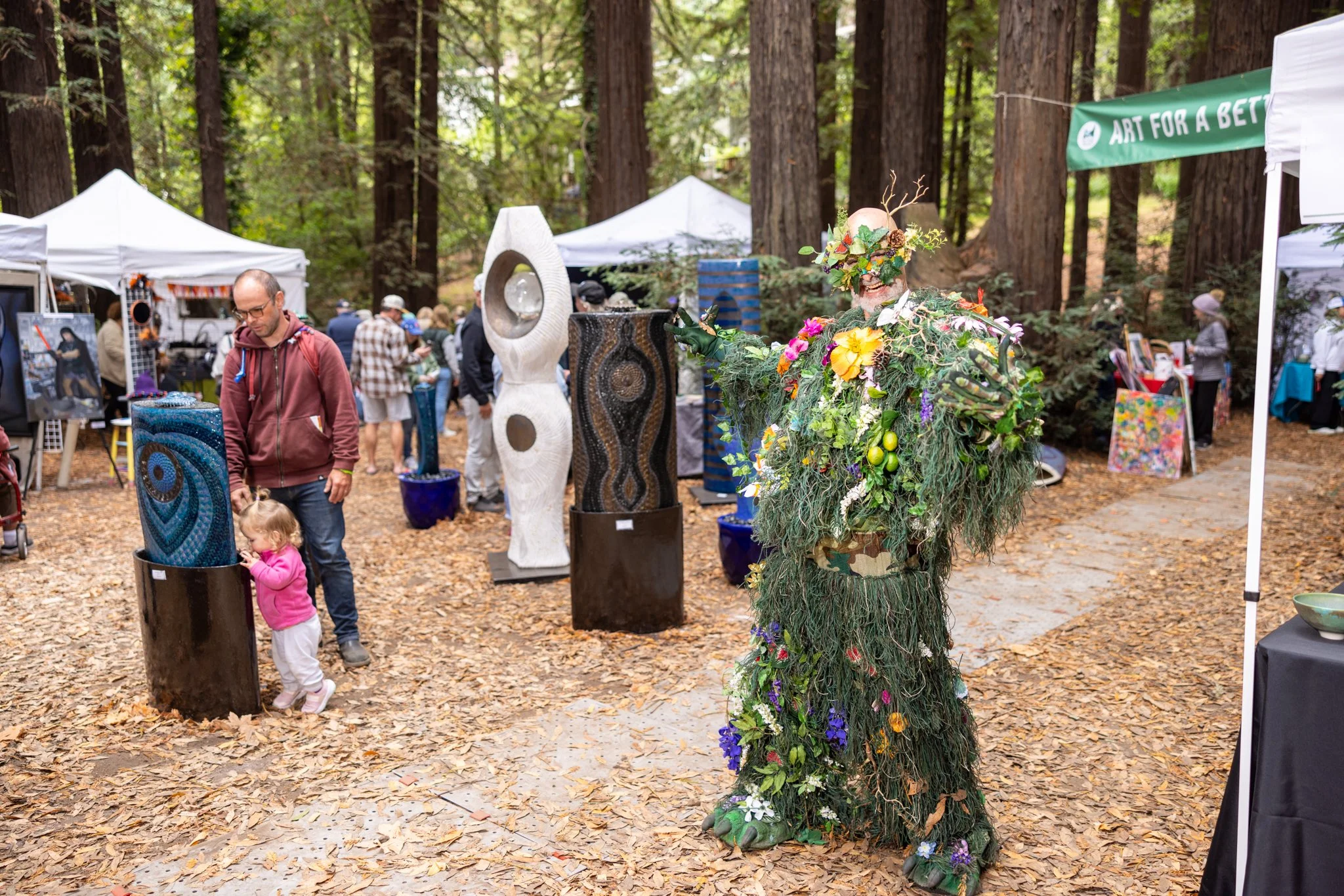 A person dressed in a costume made of greenery and flowers at an outdoor art festival with various sculptures and tents in the background.