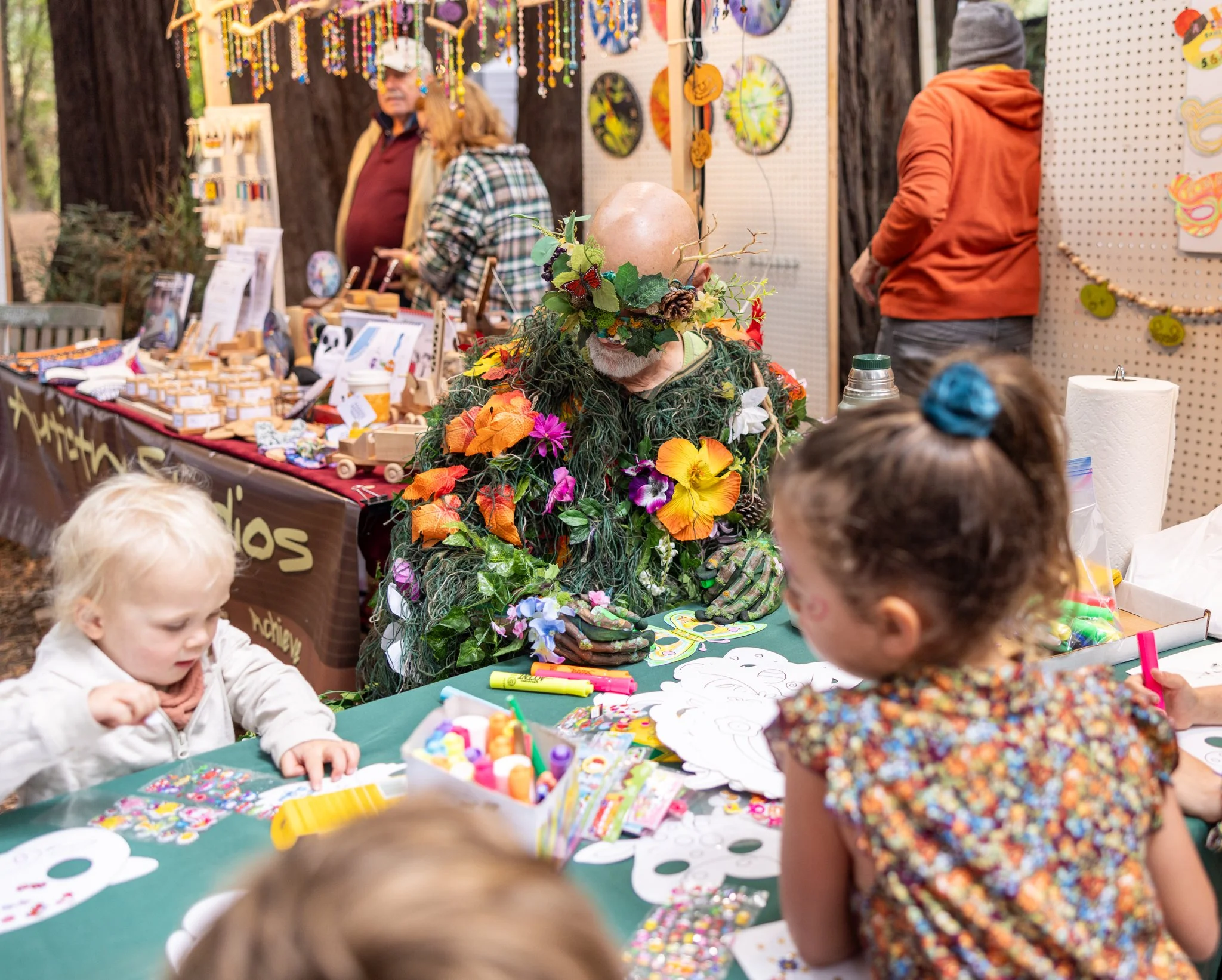 A group of children sits at a table outdoors, coloring and doing arts and crafts, while a person dressed in a colorful, nature-inspired costume with flowers, leaves, and a large head is in the background at what appears to be a craft fair or festival