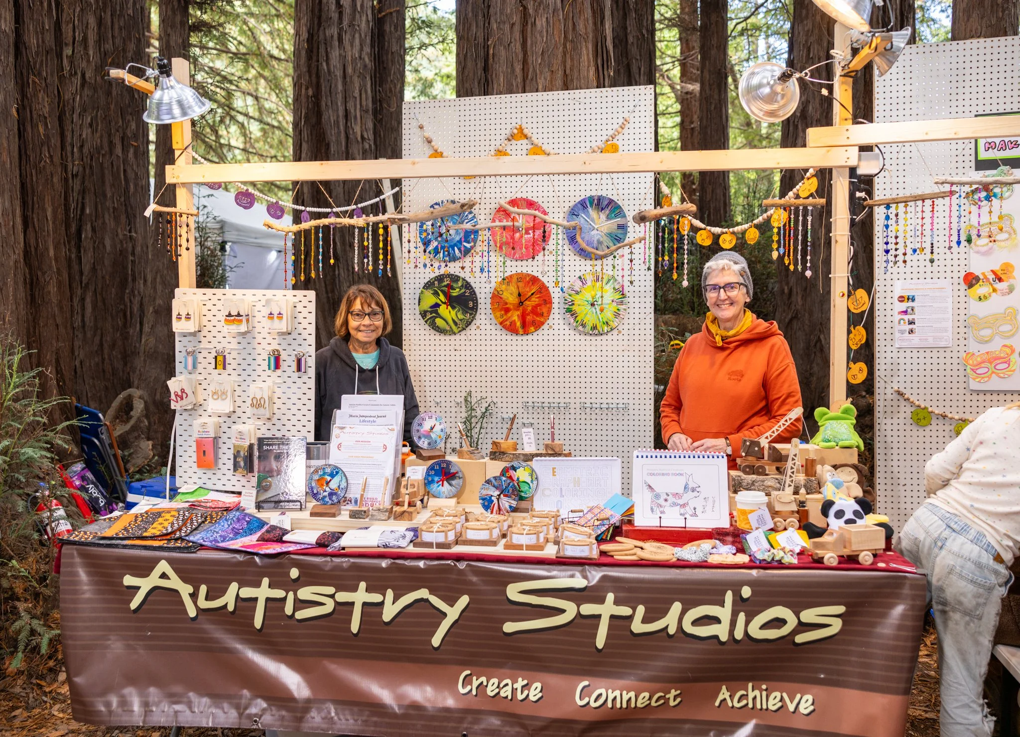 Two women standing behind a table at an outdoor craft booth called Autistry Studios, displaying handmade jewelry, clocks, wooden toys, and other craft items in a forest setting.
