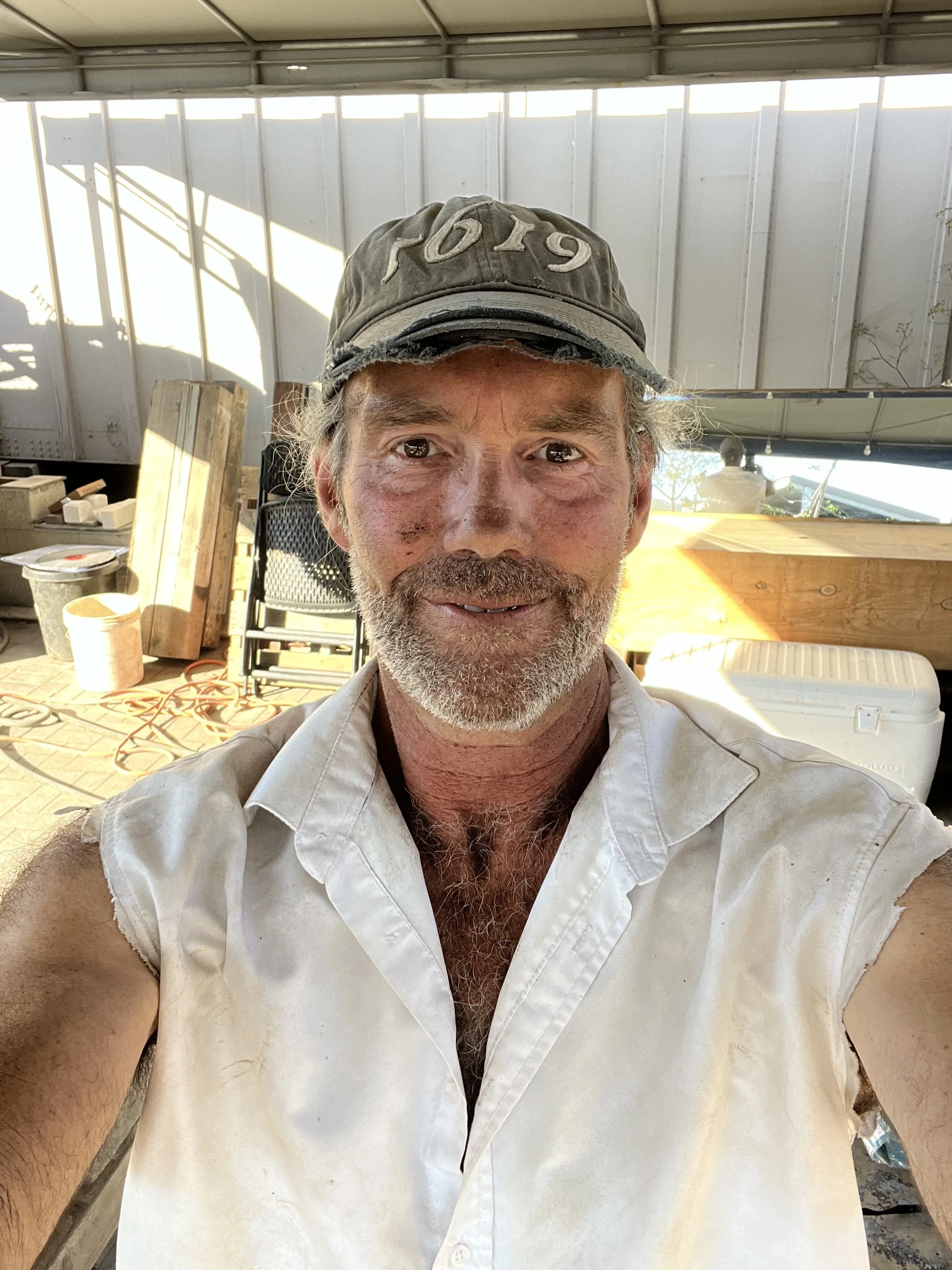 A man with a beard and mustache taking a selfie in a workshop, wearing a gray cap and a sleeveless white shirt, with woodworking tools and supplies visible in the background.