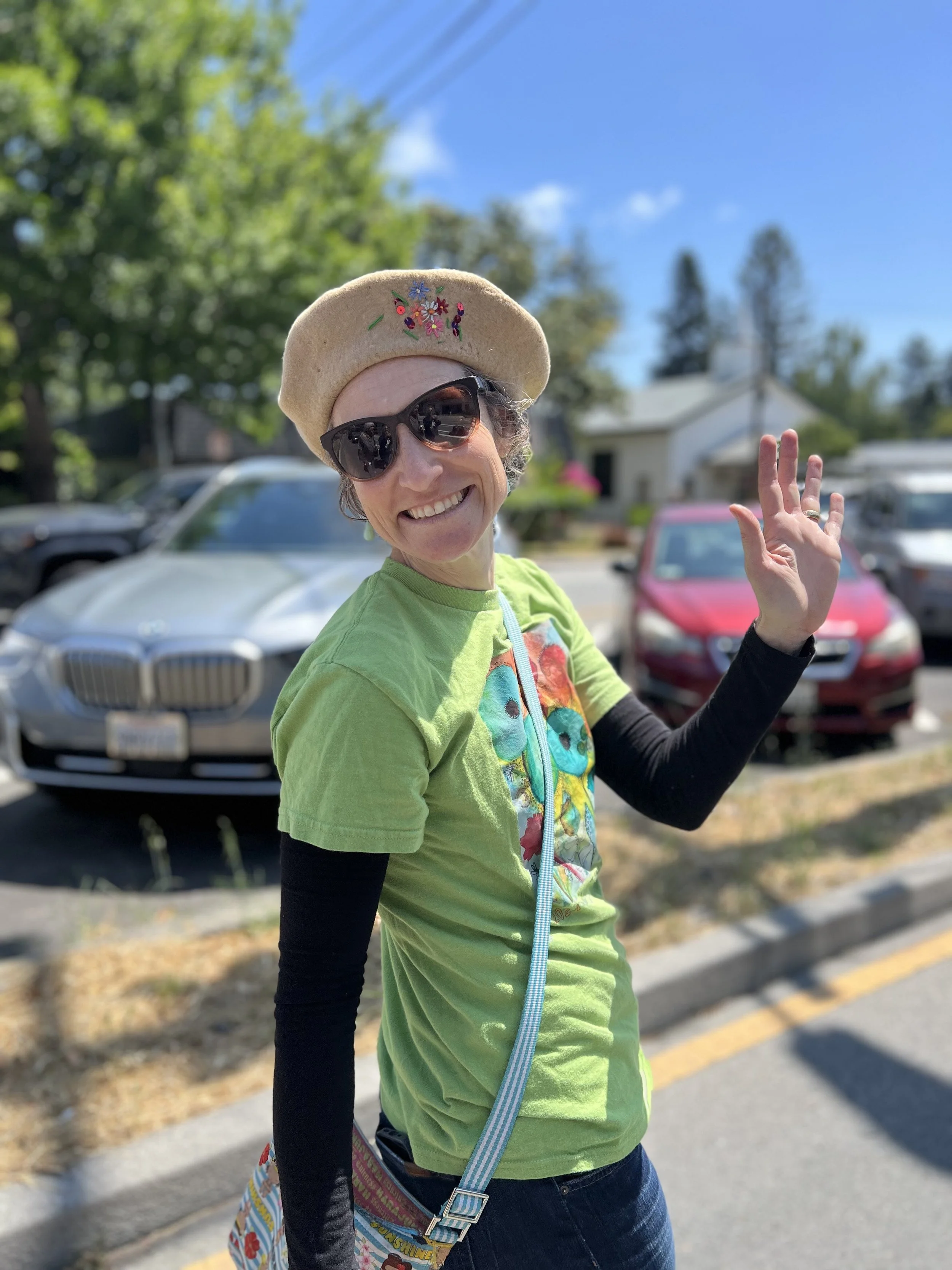 Smiling woman wearing black sunglasses, a beige beret with colorful embroidery, and a bright green t-shirt with a colorful graphic, waving outdoors on a sunny day with parked cars and trees in the background.