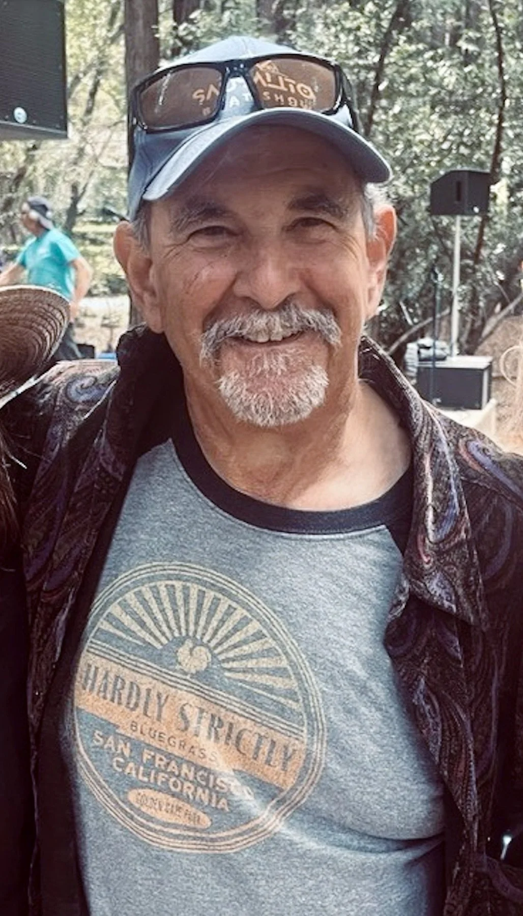 Smiling man wearing a gray baseball cap with sunglasses on top, a light gray T-shirt with a graphic design, and a dark patterned shirt open over it. The background shows trees and other people outdoors.