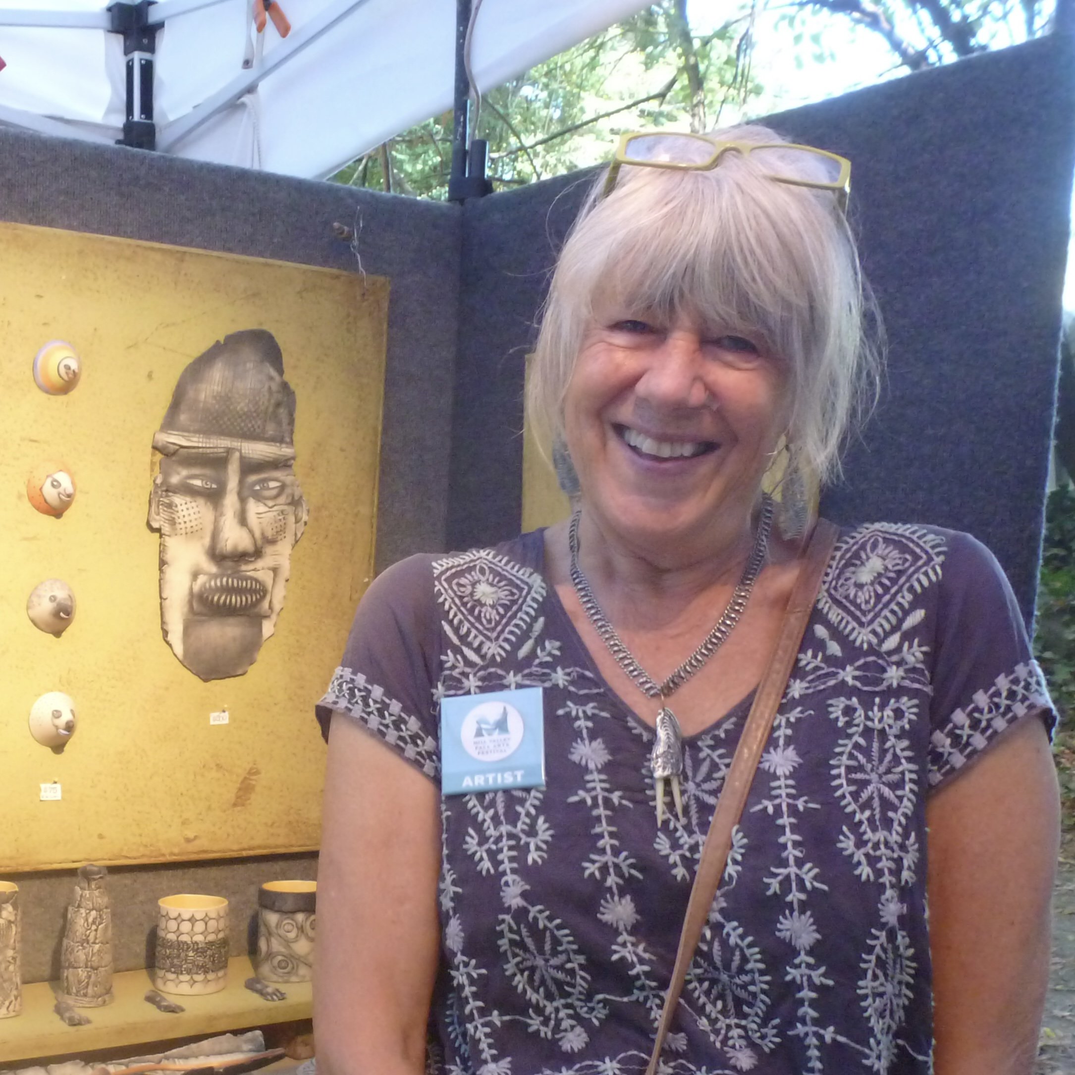 Smiling woman with glasses on her head wearing a dark embroidered shirt and an artist badge, standing in front of her art display at an outdoor event.