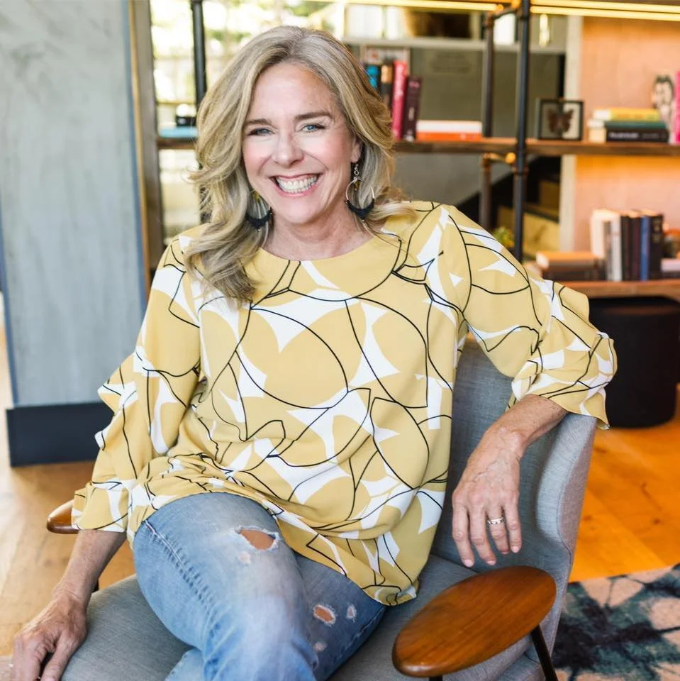 A woman with blue eyes and blond hair in a messy bun, wearing a denim jacket, sitting on a yellow chair, holding a glass of red wine, smiling indoors with wooden shelves and clothing in the background.
