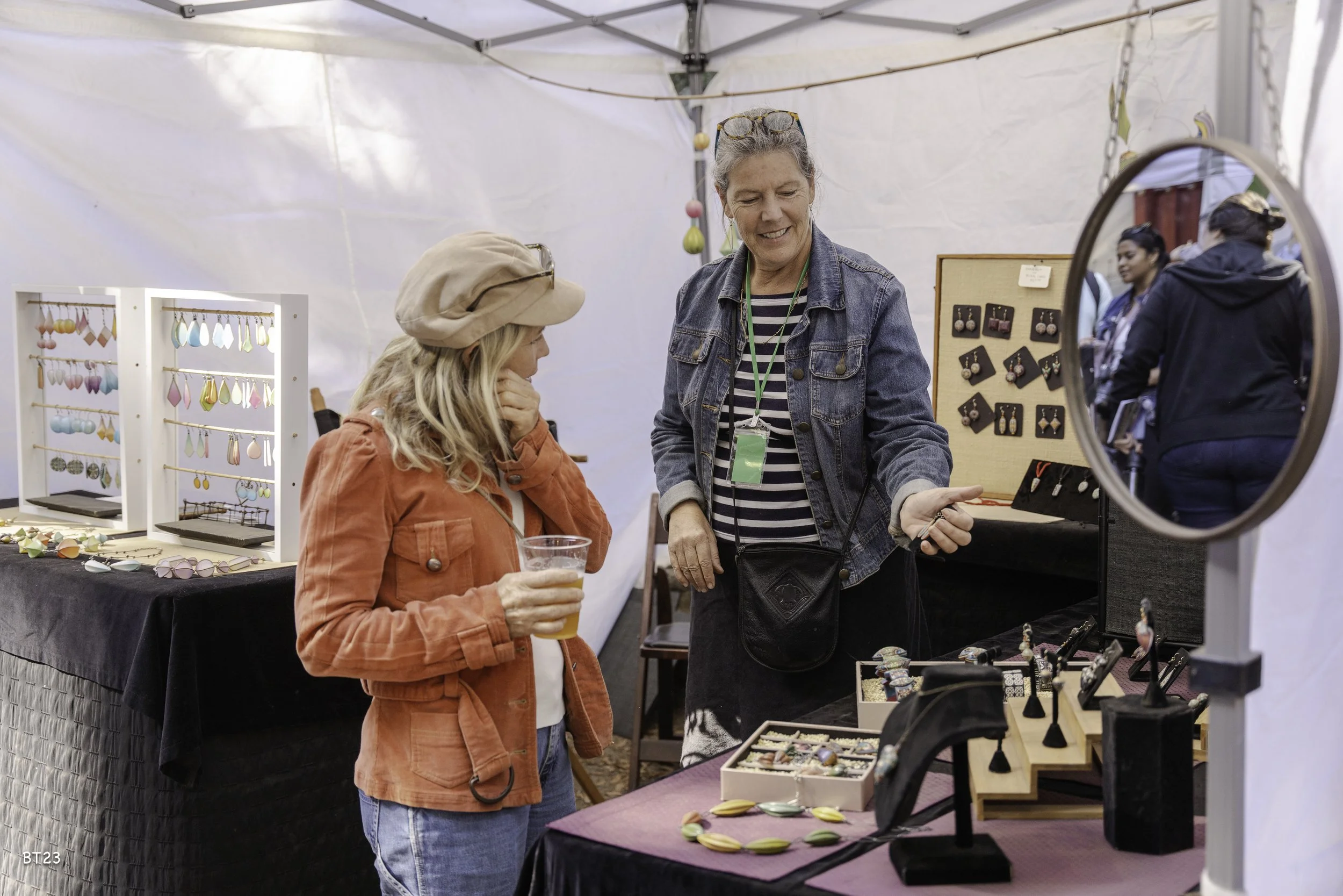 Two women at a jewelry booth, one with a drink, talking in a white tent with jewelry displays, including earrings, rings, and necklaces.