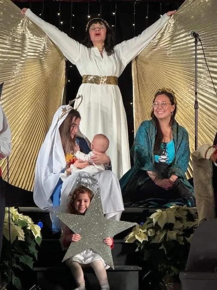 A woman dressed as Mary holding a baby Jesus doll, surrounded by women and a young girl holding a large glittery star, in a nativity scene setup during Christmas.