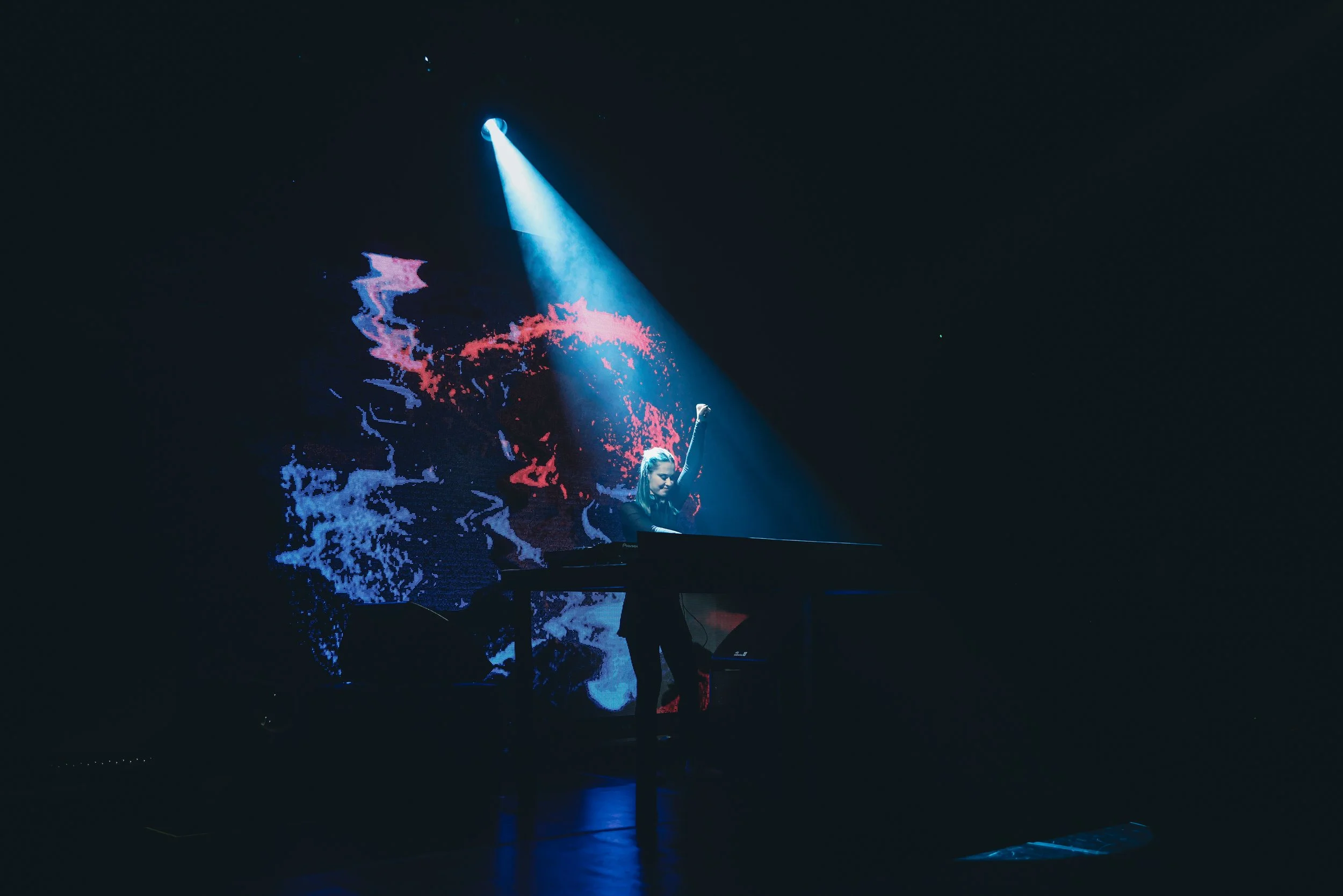 A DJ performs on stage under a bright spotlight, with a colorful abstract digital backdrop behind her, in a dark venue.
