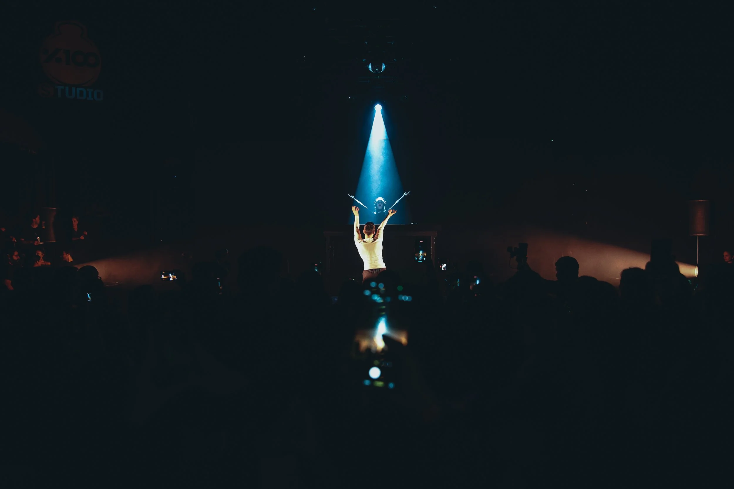 A performer with arms raised on stage, illuminated by a spotlight, with an audience watching and capturing the moment on their phones.