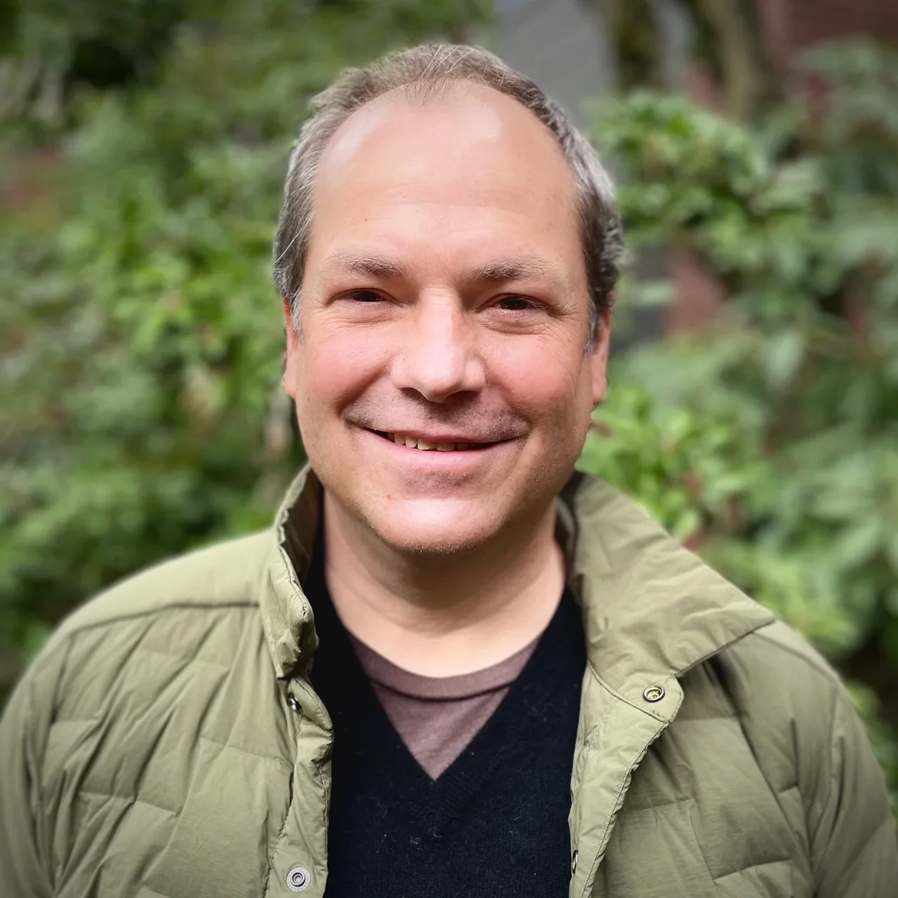 A man smiling outdoors with green foliage in the background.