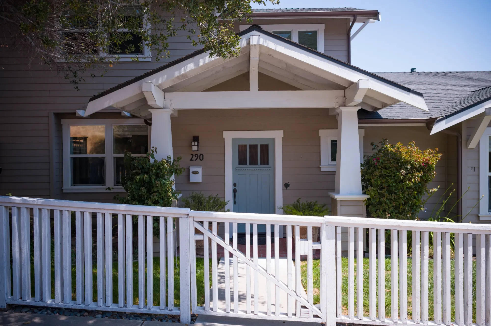 Front view of a beige house with white trim, featuring a small porch with white posts and a light blue door, surrounded by a white picket fence and greenery.