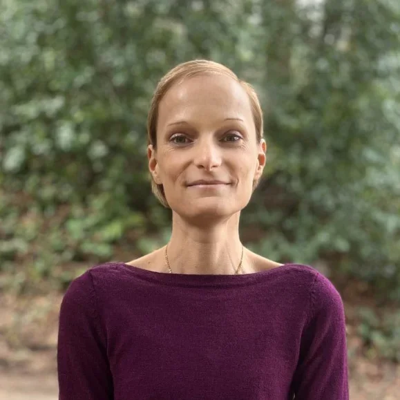 A woman with short blonde hair, wearing a purple top, standing outdoors with greenery in the background.