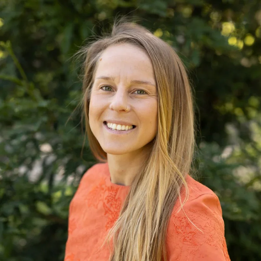 Smiling woman with long, light brown hair and a nose piercing, wearing an orange top, outdoors with green foliage background.