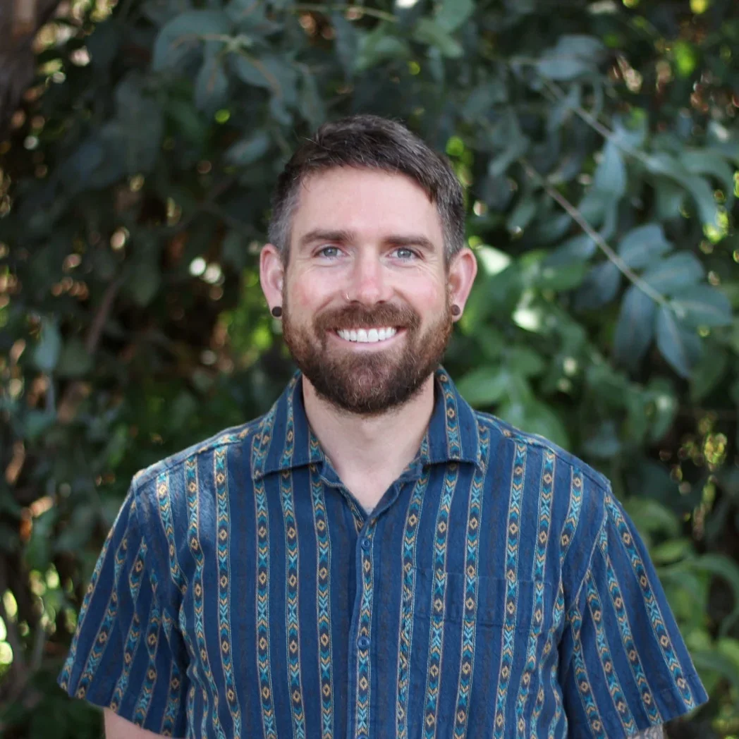 A smiling man with a beard, short dark hair, and earrings, standing outdoors in front of green foliage, wearing a patterned blue shirt.
