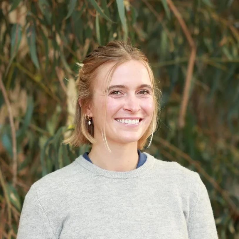 A young woman with blonde hair smiling outdoors, wearing a light gray sweater and earrings.