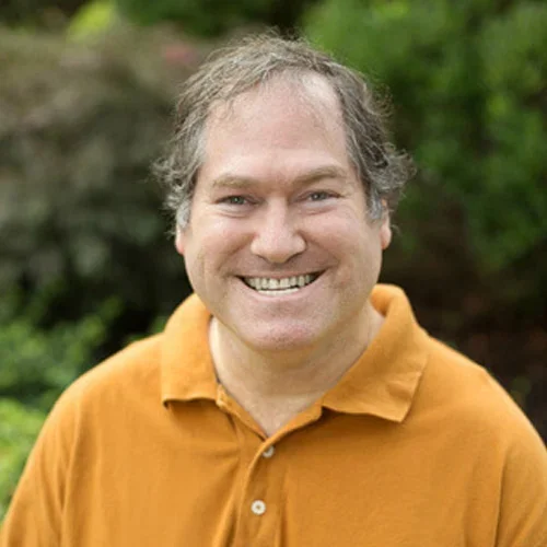 A smiling man with curly gray hair wearing an orange polo shirt outdoors with green foliage in the background.