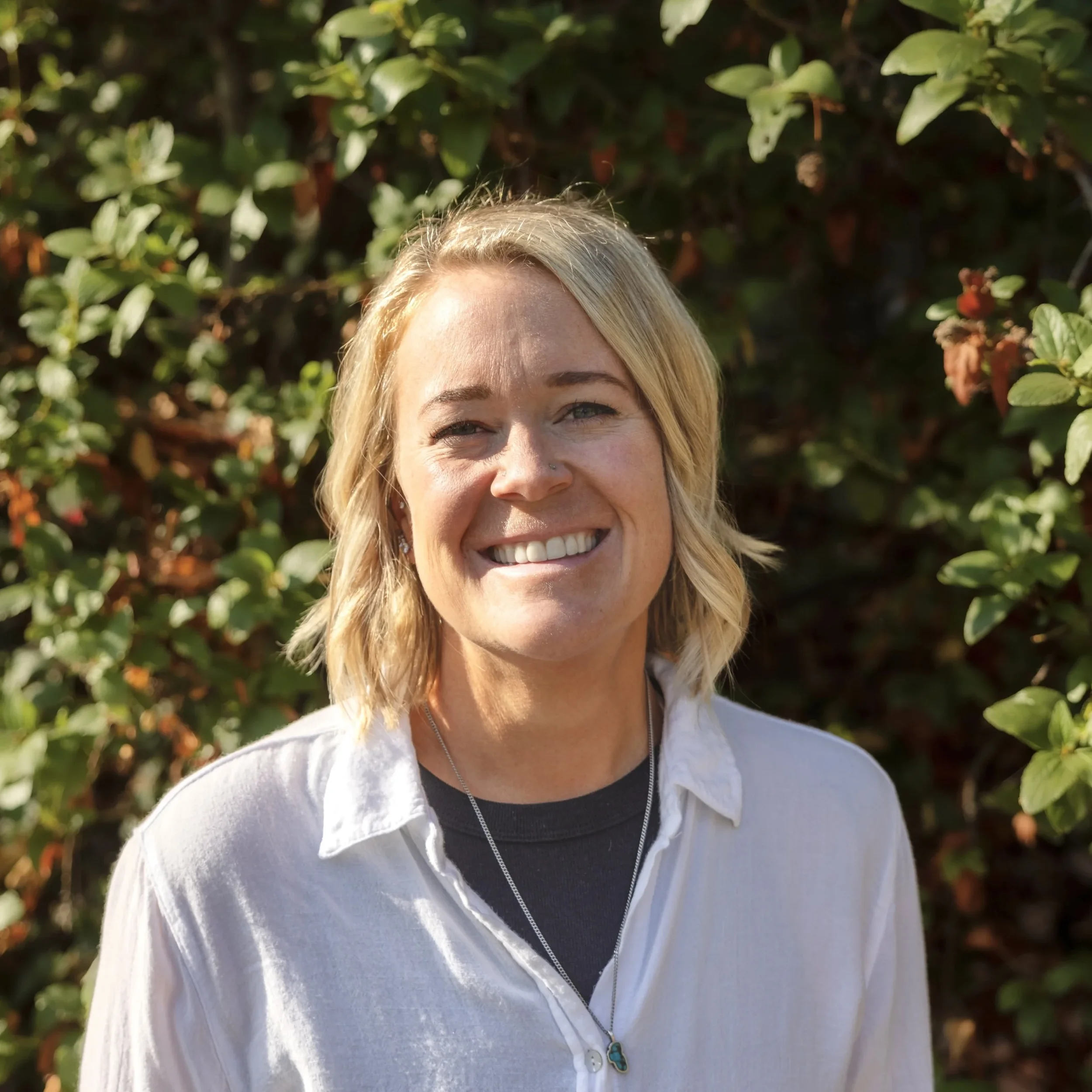 A woman with blonde hair smiling outdoors in front of green foliage, wearing a white shirt and a necklace.