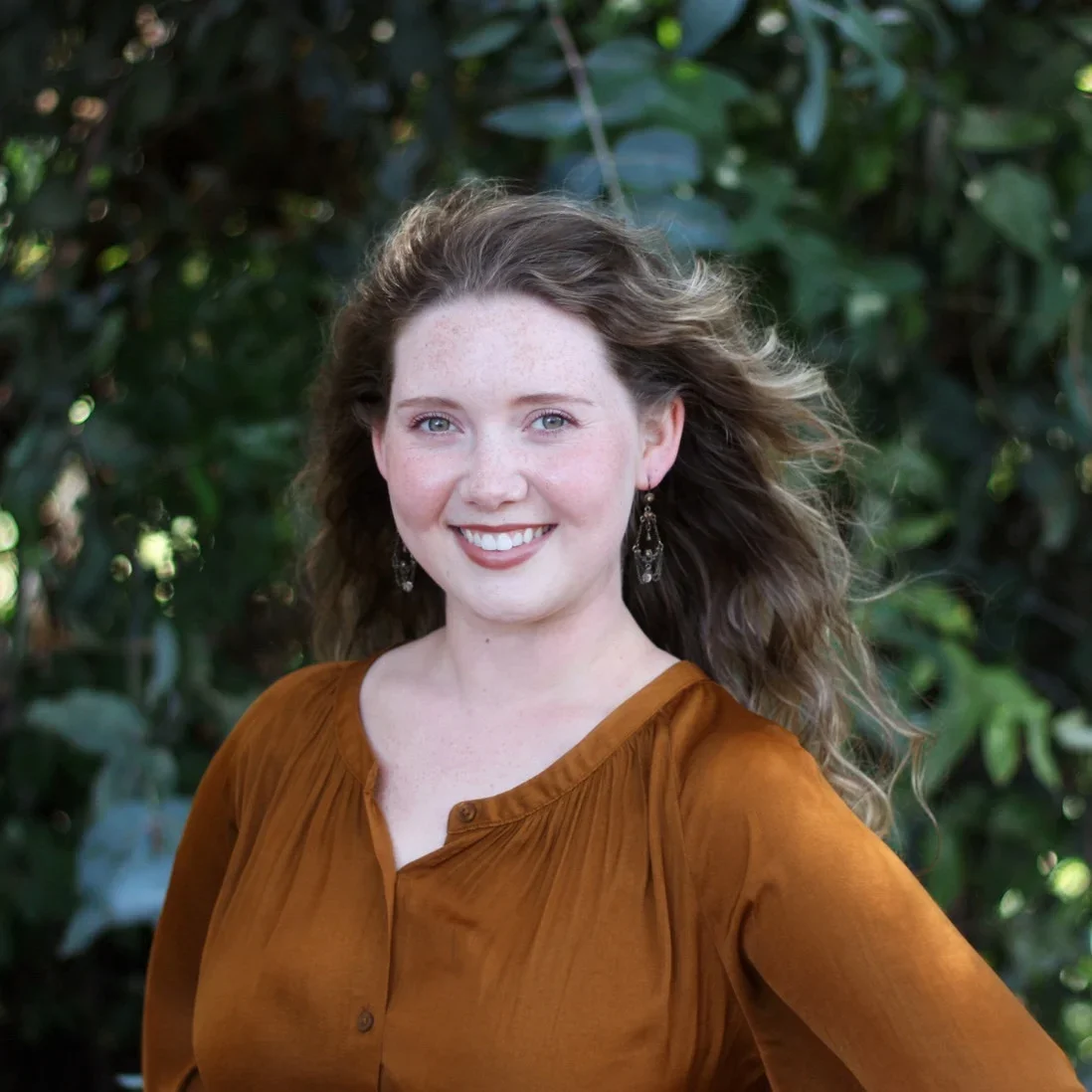 A young woman with wavy brown hair, light skin, and blue eyes, smiling outdoors with greenery in the background, wearing a rust-colored top and dangling earrings.