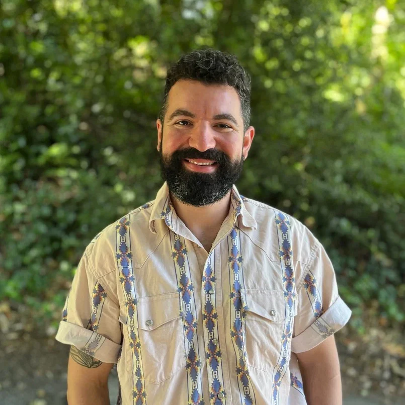 A man with dark hair and a full beard smiling outdoors with a background of green trees and foliage.