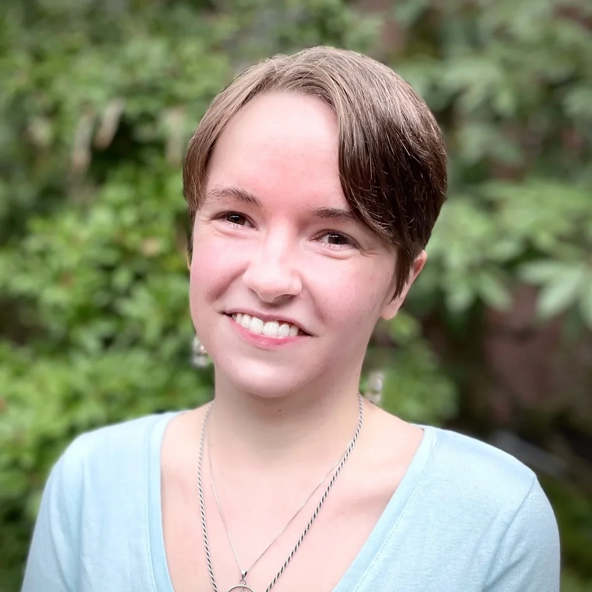 A smiling young person with short brown hair wearing a light blue shirt and layered necklaces outdoors with greenery in the background.