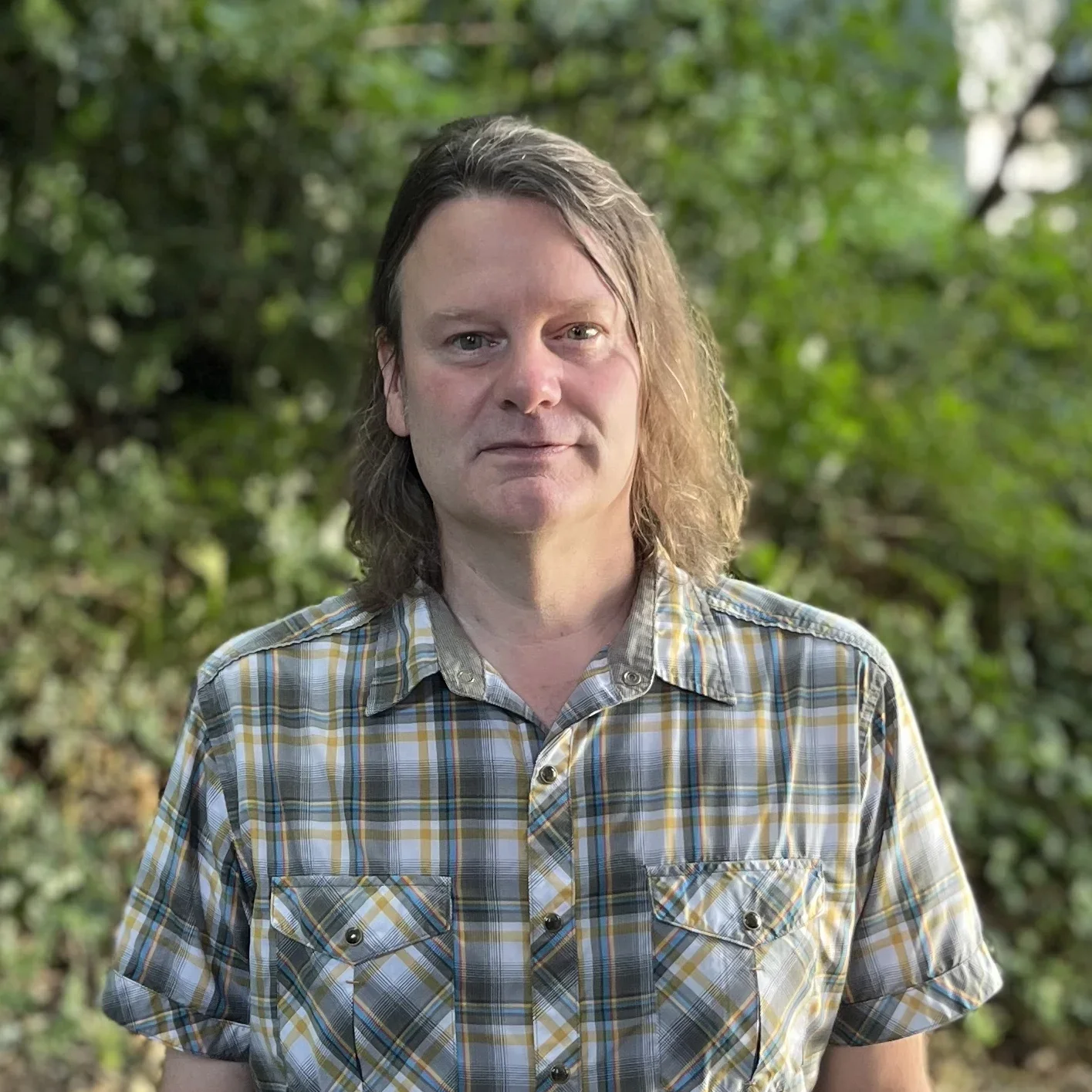 A man with shoulder-length brown hair, wearing a yellow plaid shirt, stands outdoors with greenery in the background.